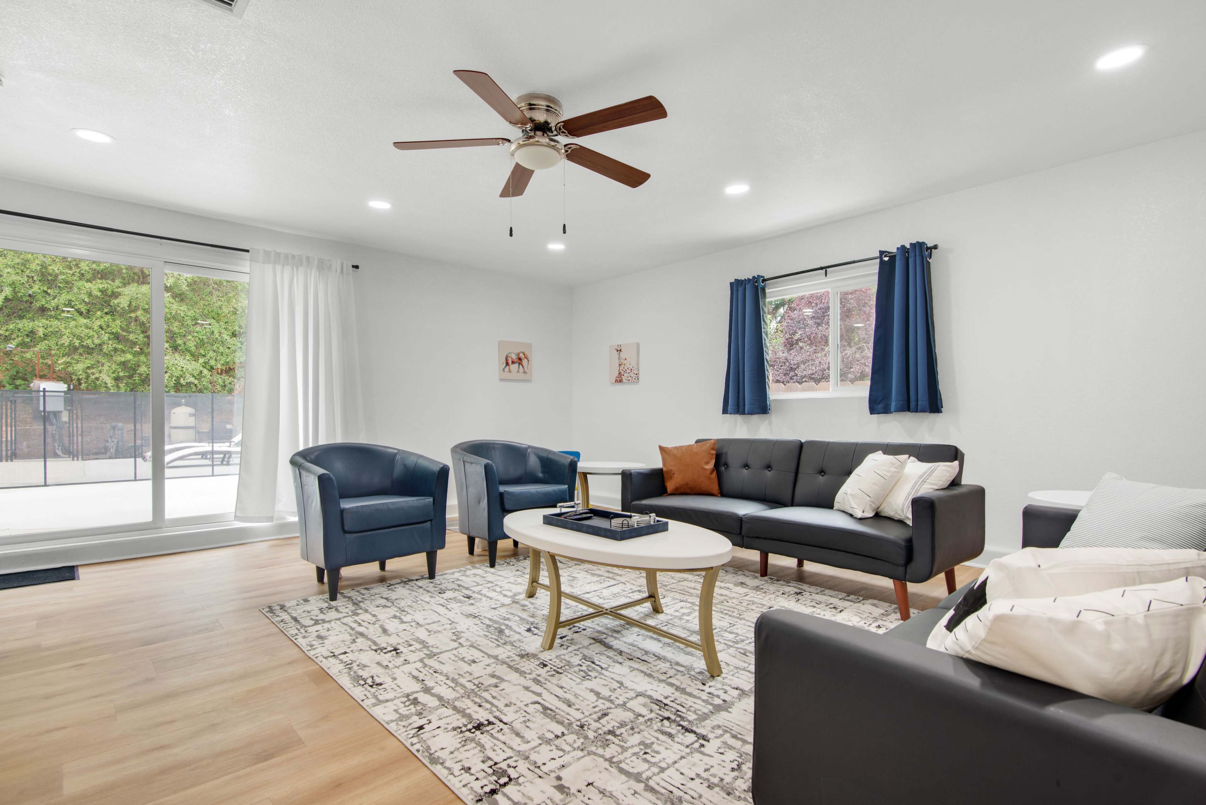 The living room features a seating area with two blue armchairs, a black sofa, a round coffee table, and a large window overlooking a patio.
