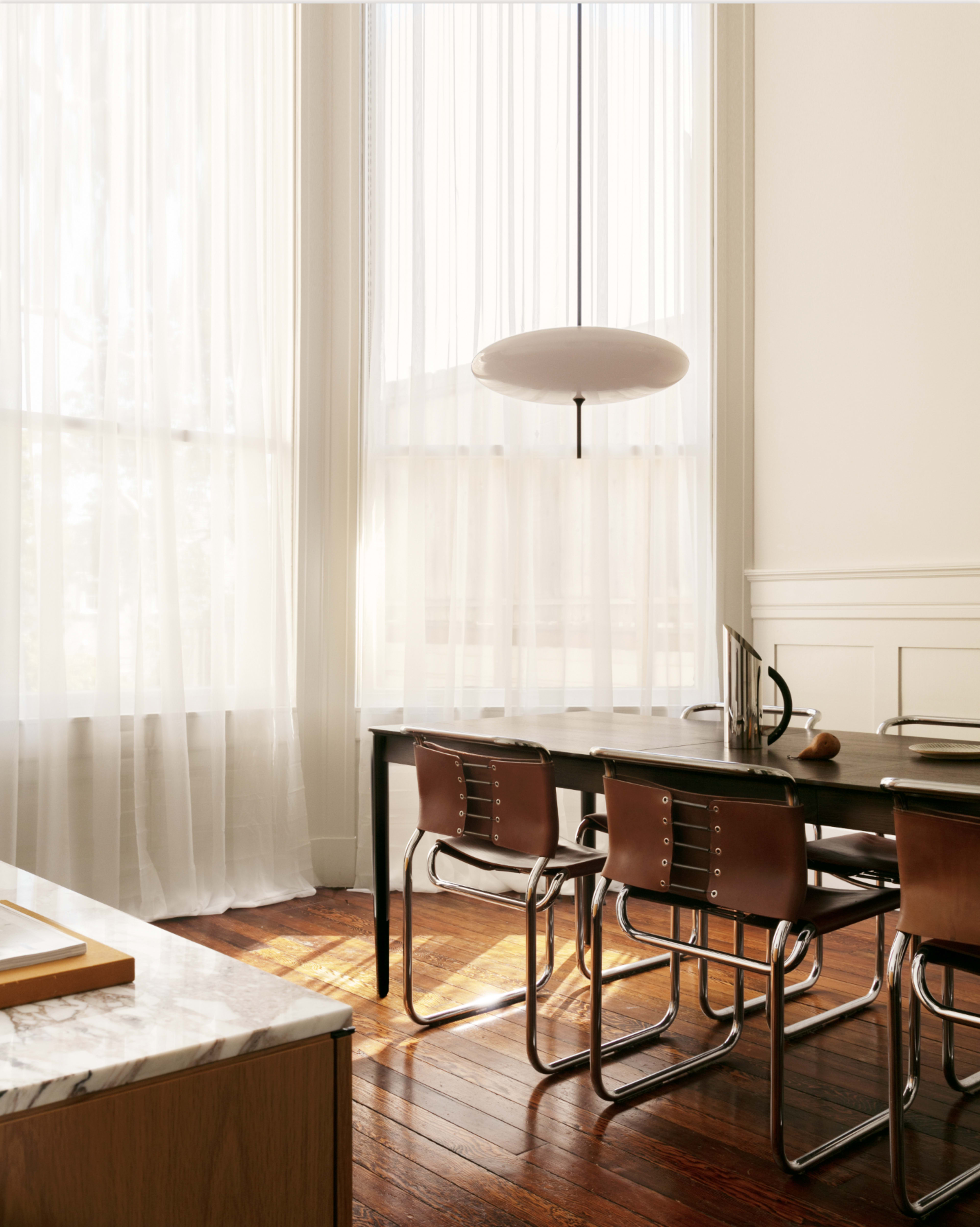 A modern dining area features a long black table surrounded by metal and leather chairs, illuminated by natural light filtering through sheer white curtains.