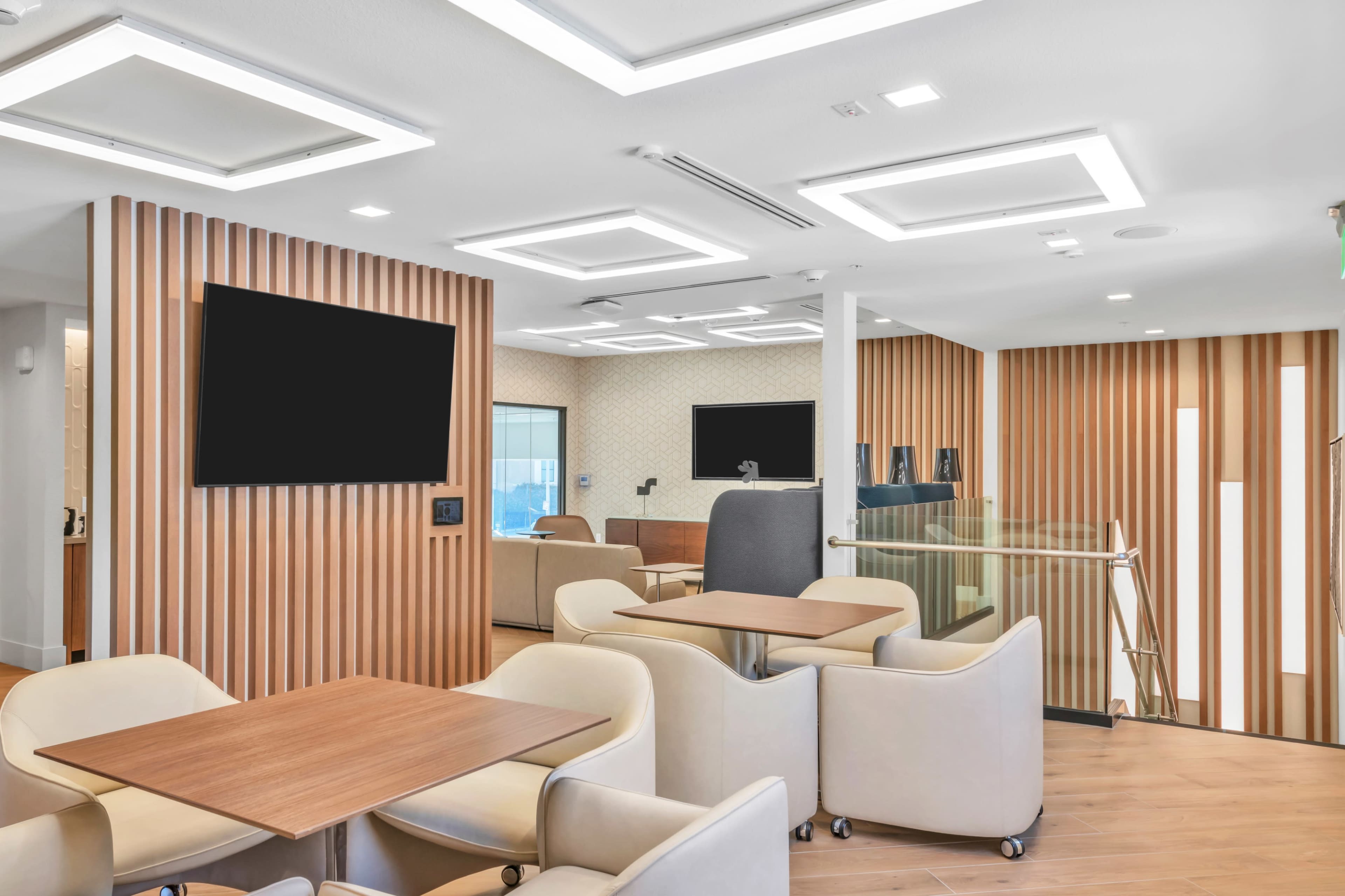 The image shows a modern open space with wooden paneling, seating areas, and two large television screens mounted on the walls.