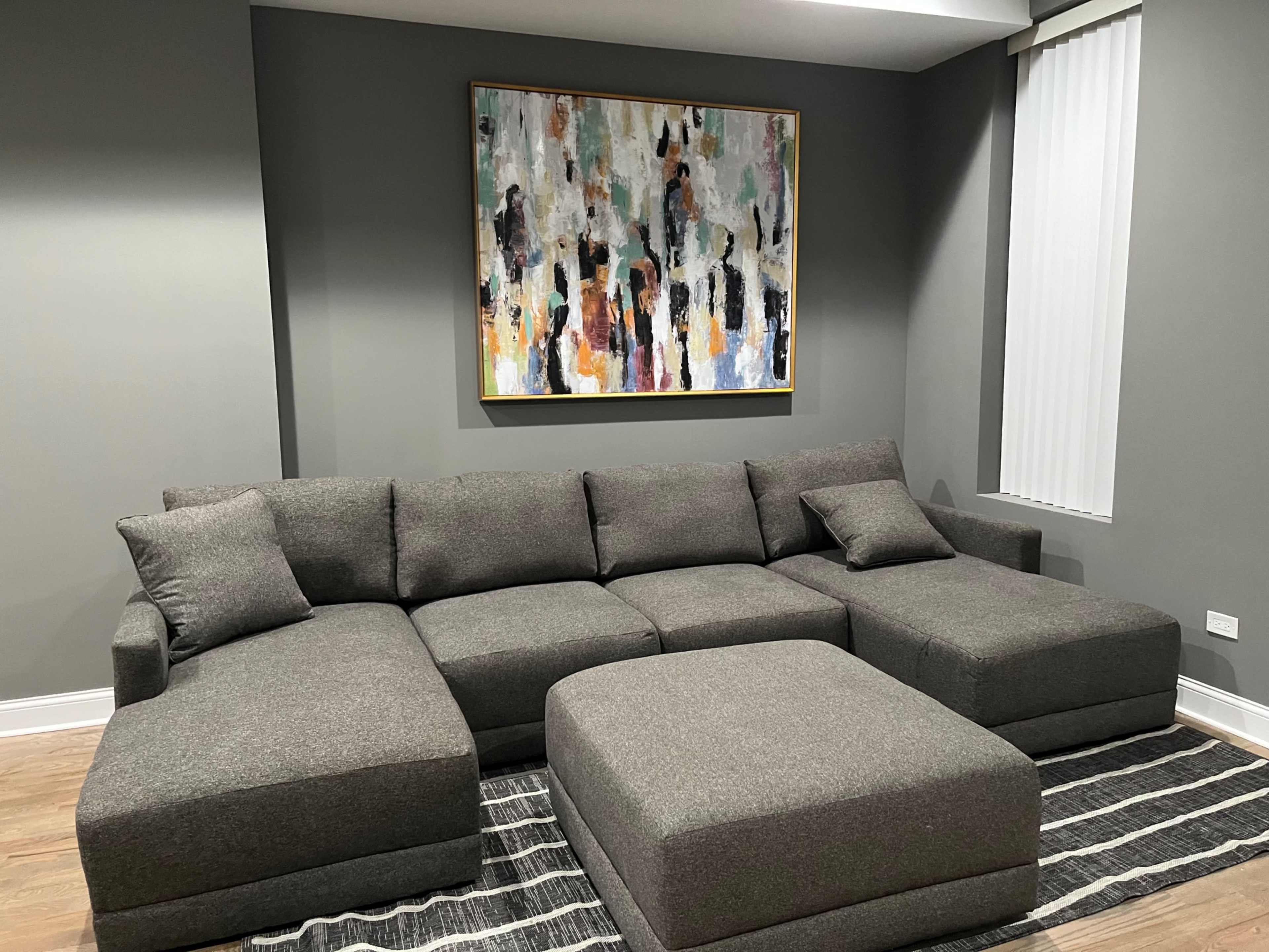 A gray sectional sofa with several cushions is positioned against a dark wall, beneath a colorful abstract painting, on a striped rug.