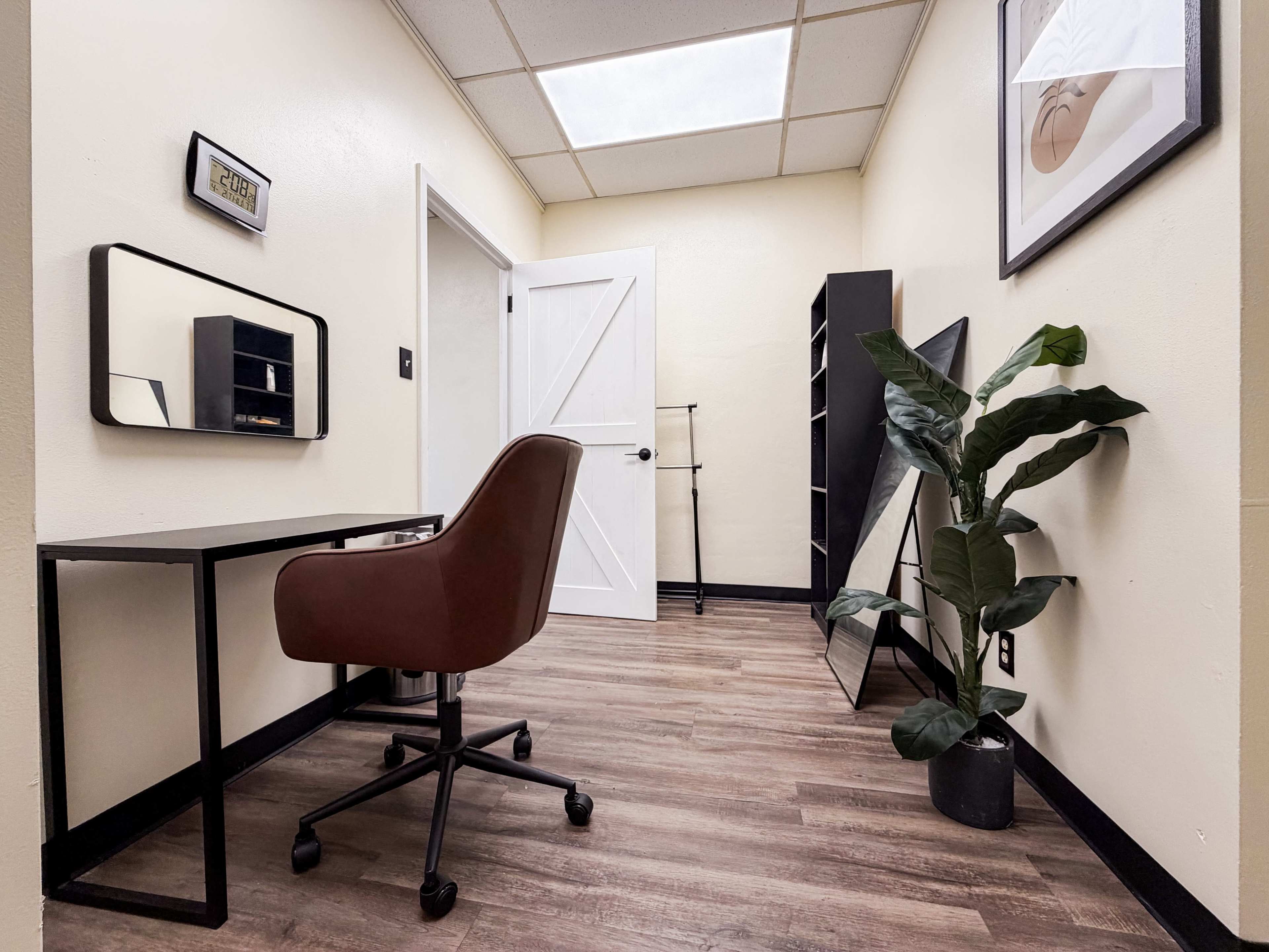 The image depicts a small office space featuring a desk with a chair, a mirror, a bookshelf, and a potted plant, all set against a beige wall.