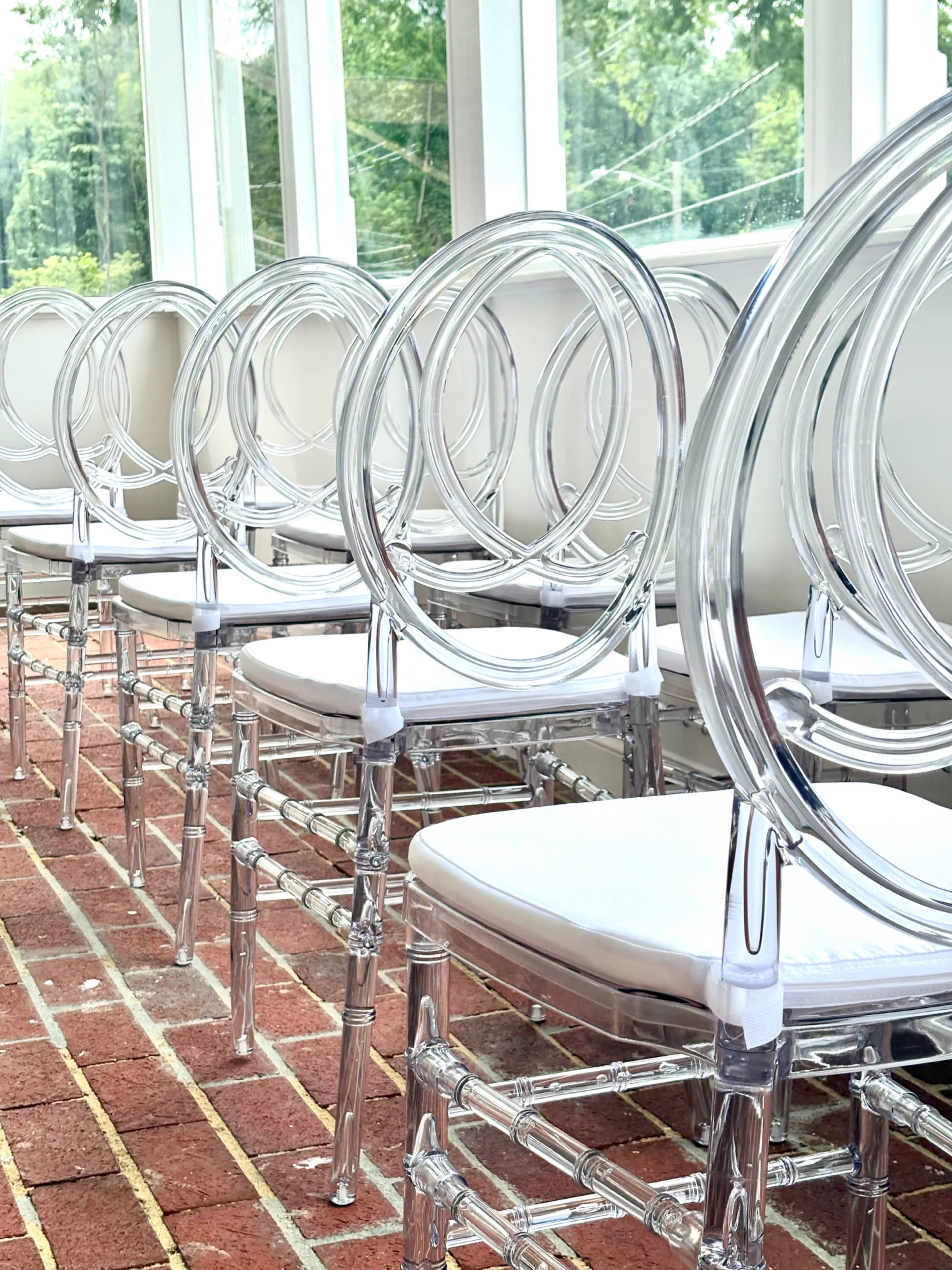 A row of clear acrylic chairs with circular backs is lined up on a brick floor in a sunlit room.
