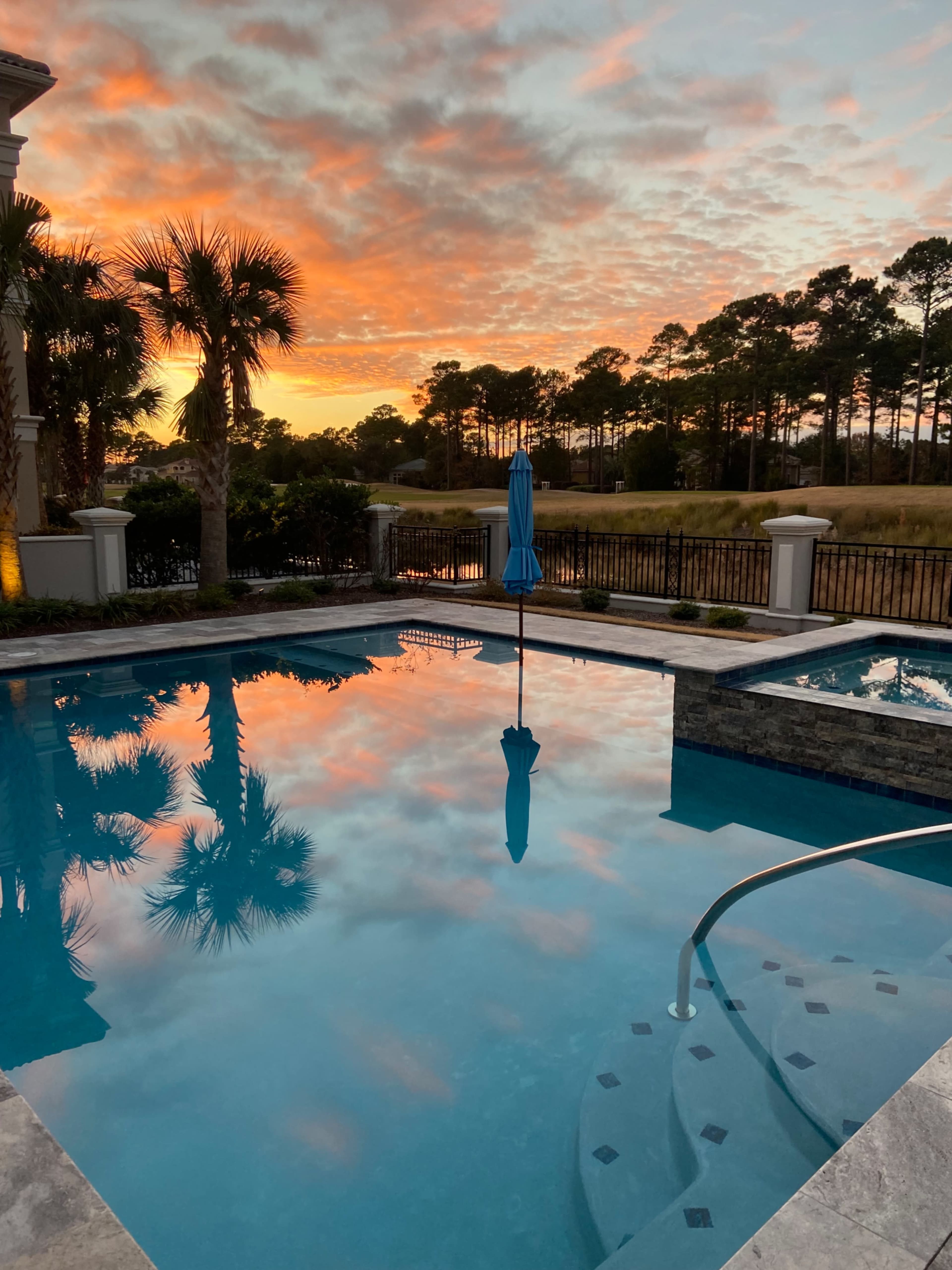 A swimming pool reflects a colorful sunset, surrounded by palm trees and a fenced area.
