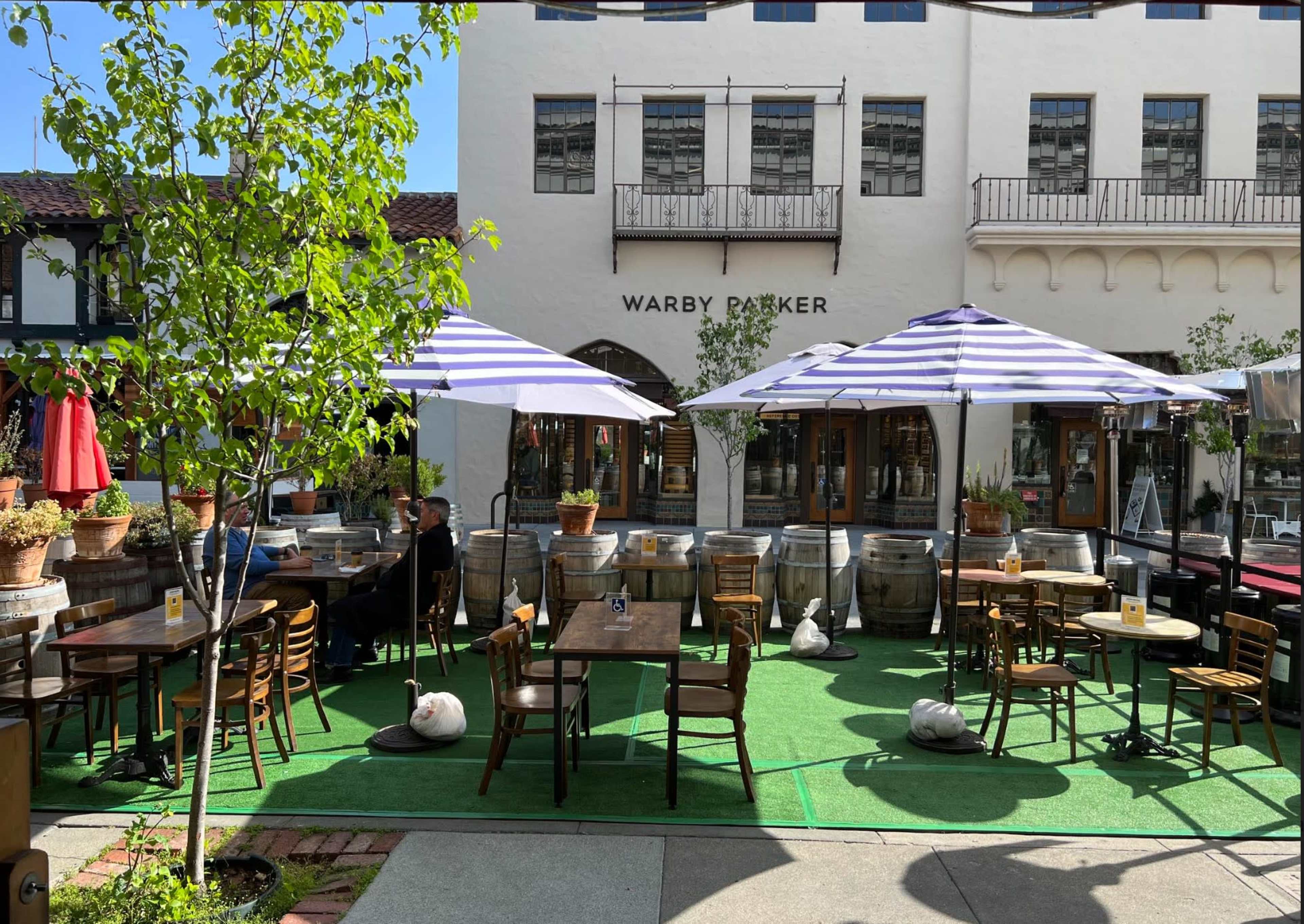 The image shows an outdoor dining area with tables and chairs under striped umbrellas, surrounded by potted plants and barrels near a storefront labeled "WARBY PARKER."