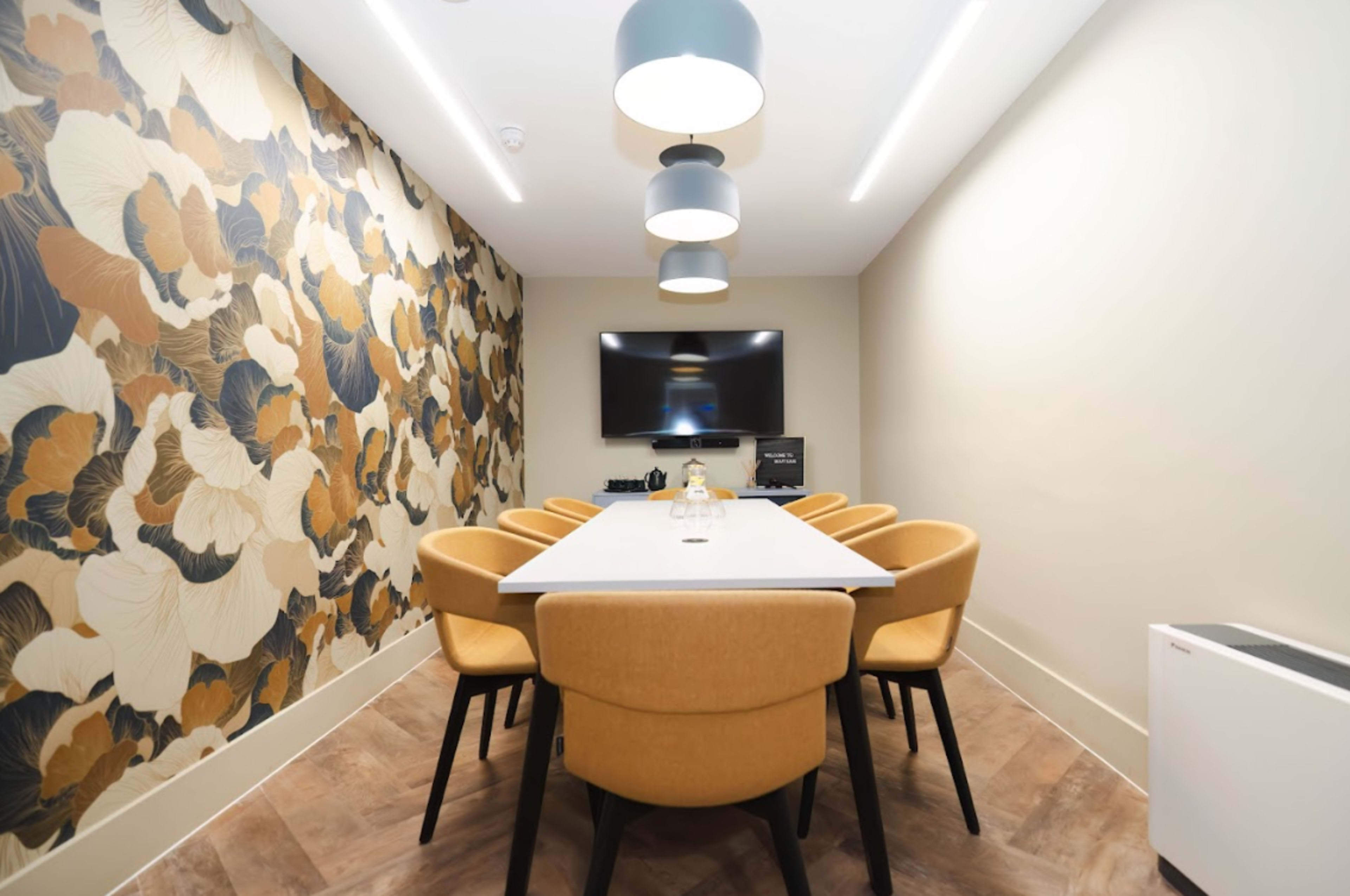 A conference room features a long white table surrounded by eight tan chairs and a decorative wall, with a television and modern lighting overhead.