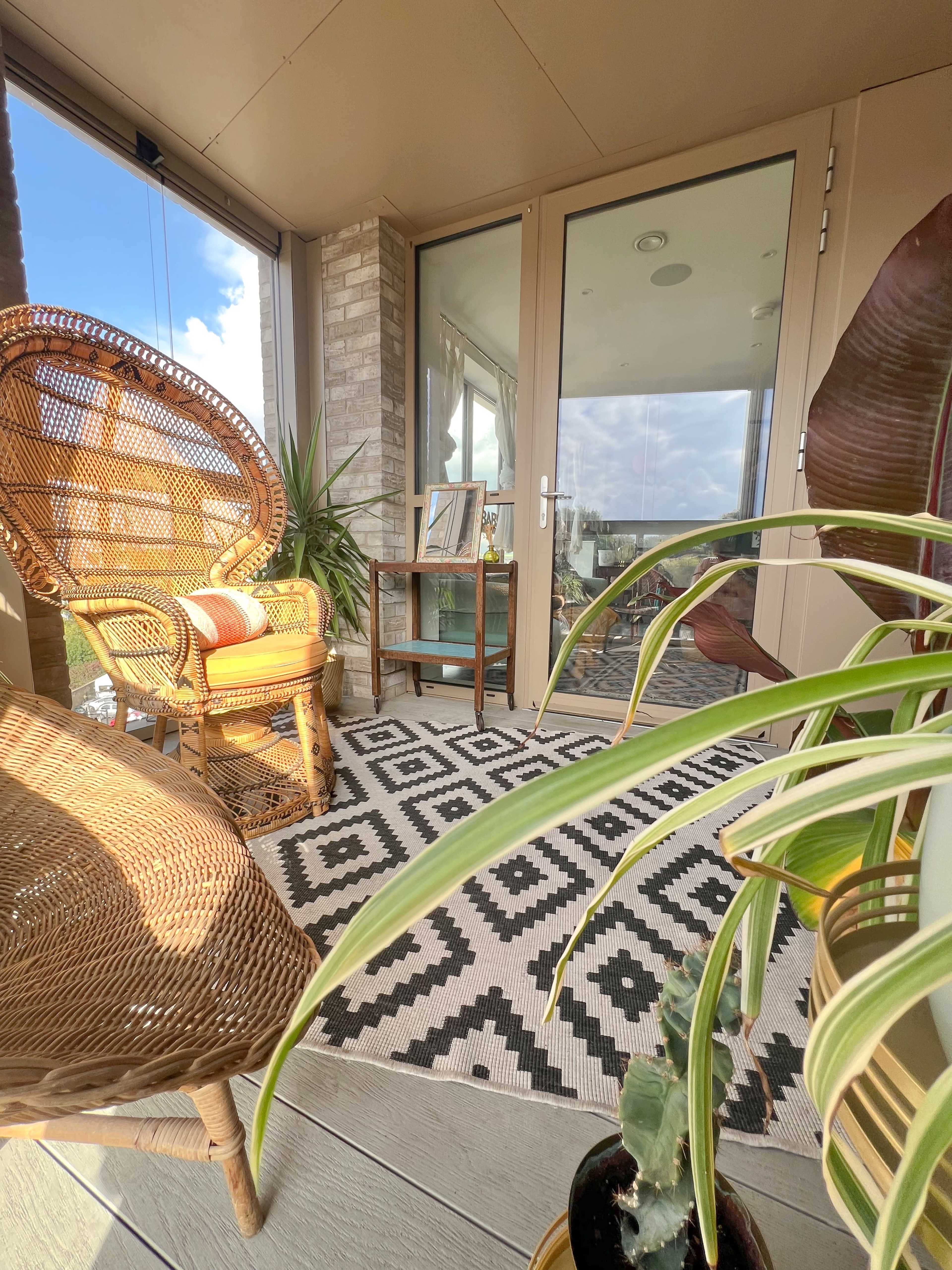A cozy outdoor terrace features two wicker chairs, a patterned rug, and potted plants near large glass doors.