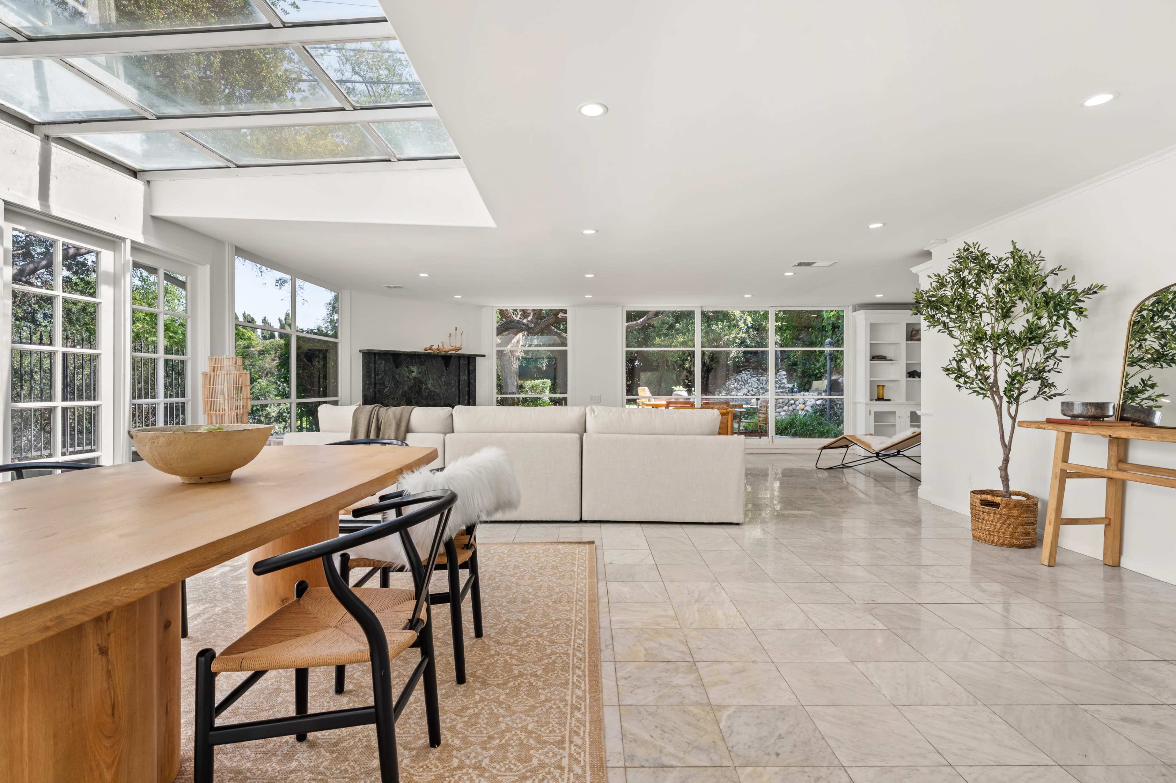 The image shows a spacious, brightly lit living area with a dining table, modern furniture, and large windows that open to a garden.