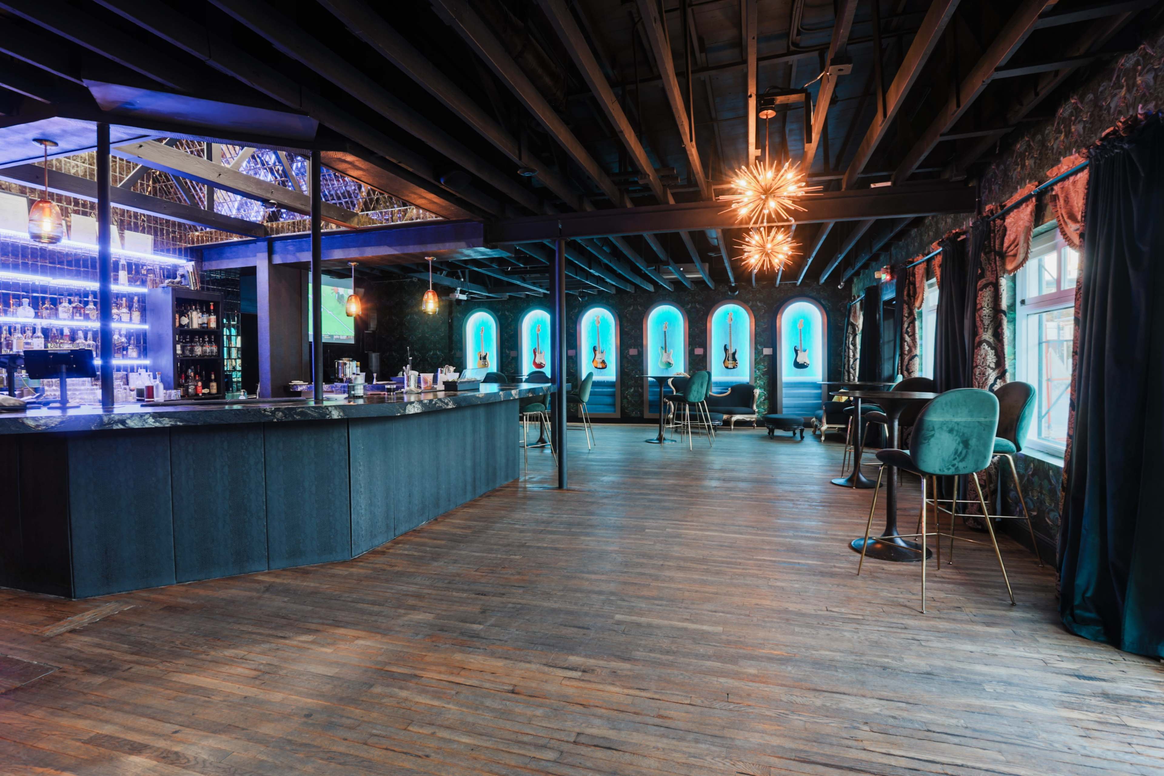 A modern bar area with a dark wooden floor, illuminated blue windows, and stylish seating around a curved counter.
