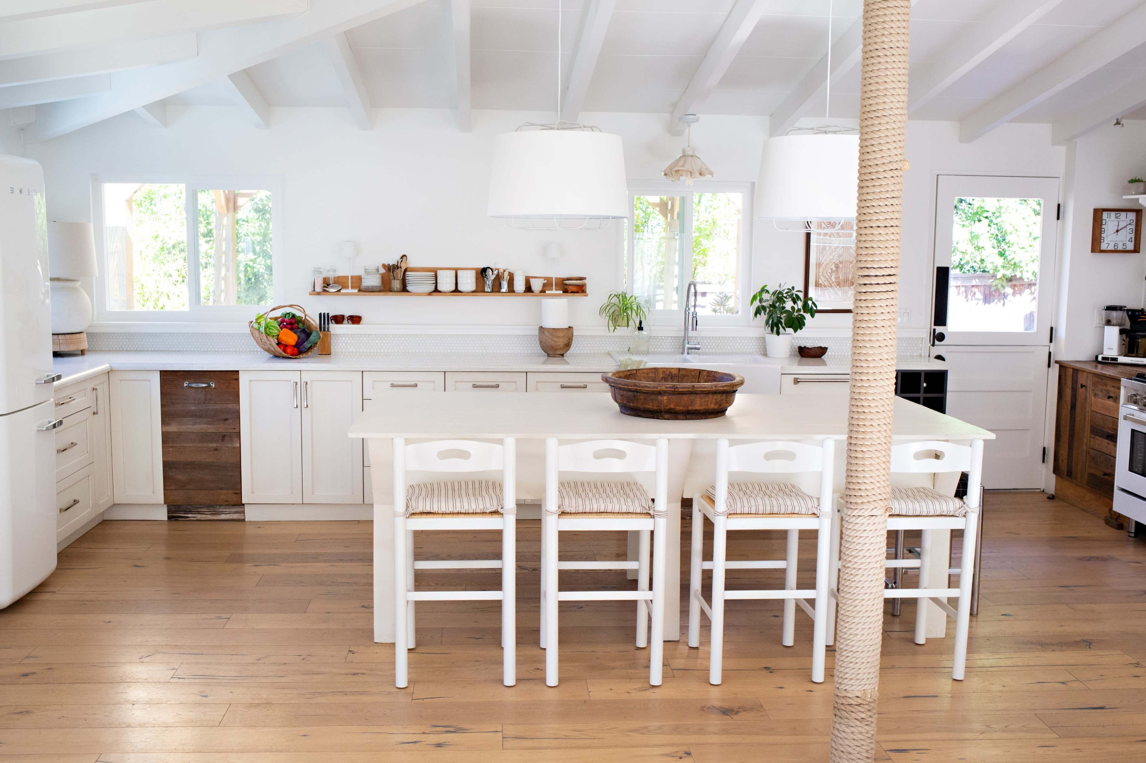 A modern kitchen features a light color scheme, a large island with white chairs, and wooden accents.