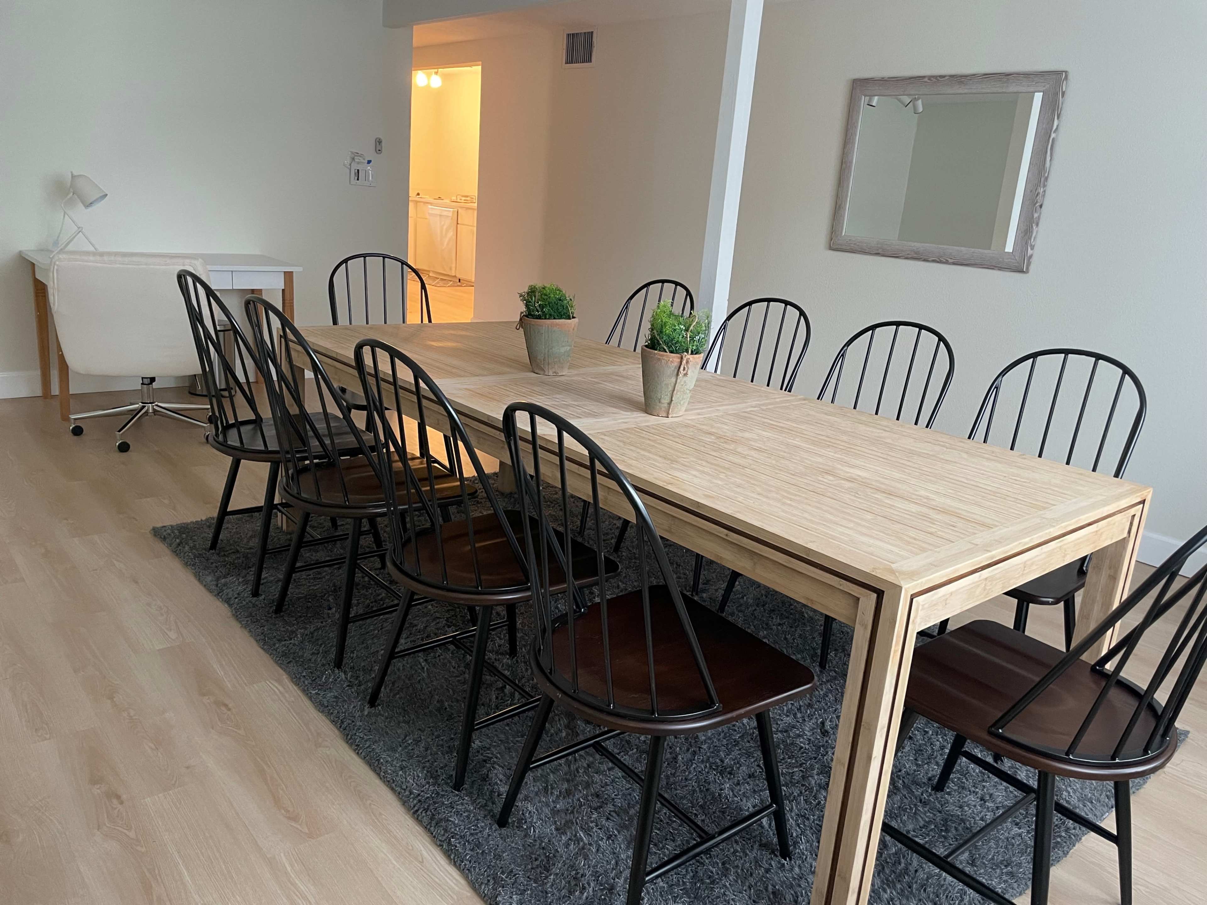A long wooden dining table with ten black chairs is set in a bright room with a gray area rug and a mirror on the wall.