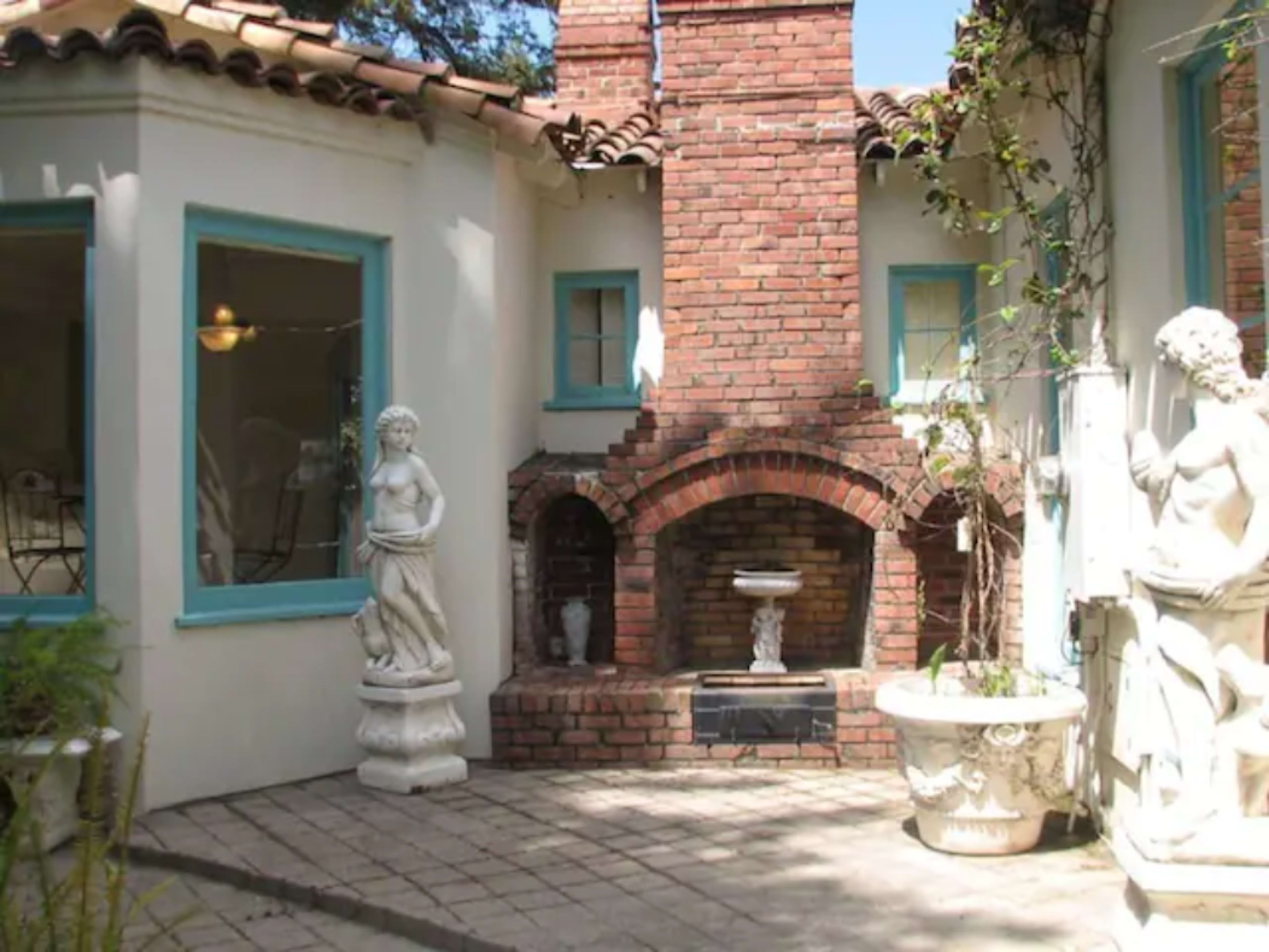A small courtyard with a brick fireplace flanked by two white statues and surrounded by a white stucco wall and blue window frames.