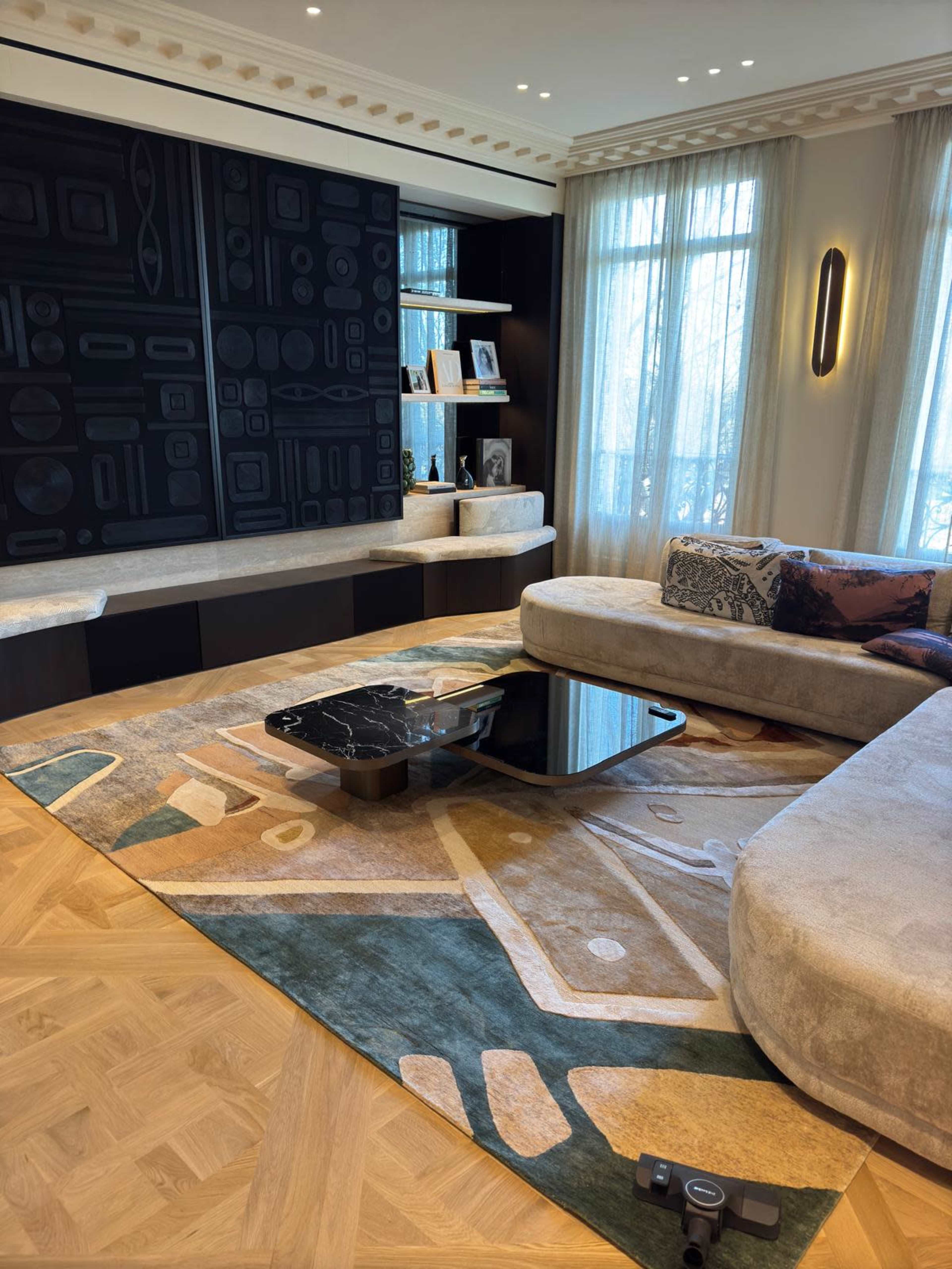 The image depicts a stylish living room featuring a curved sofa, a low black coffee table, a patterned area rug, and a large black wall with geometric designs.