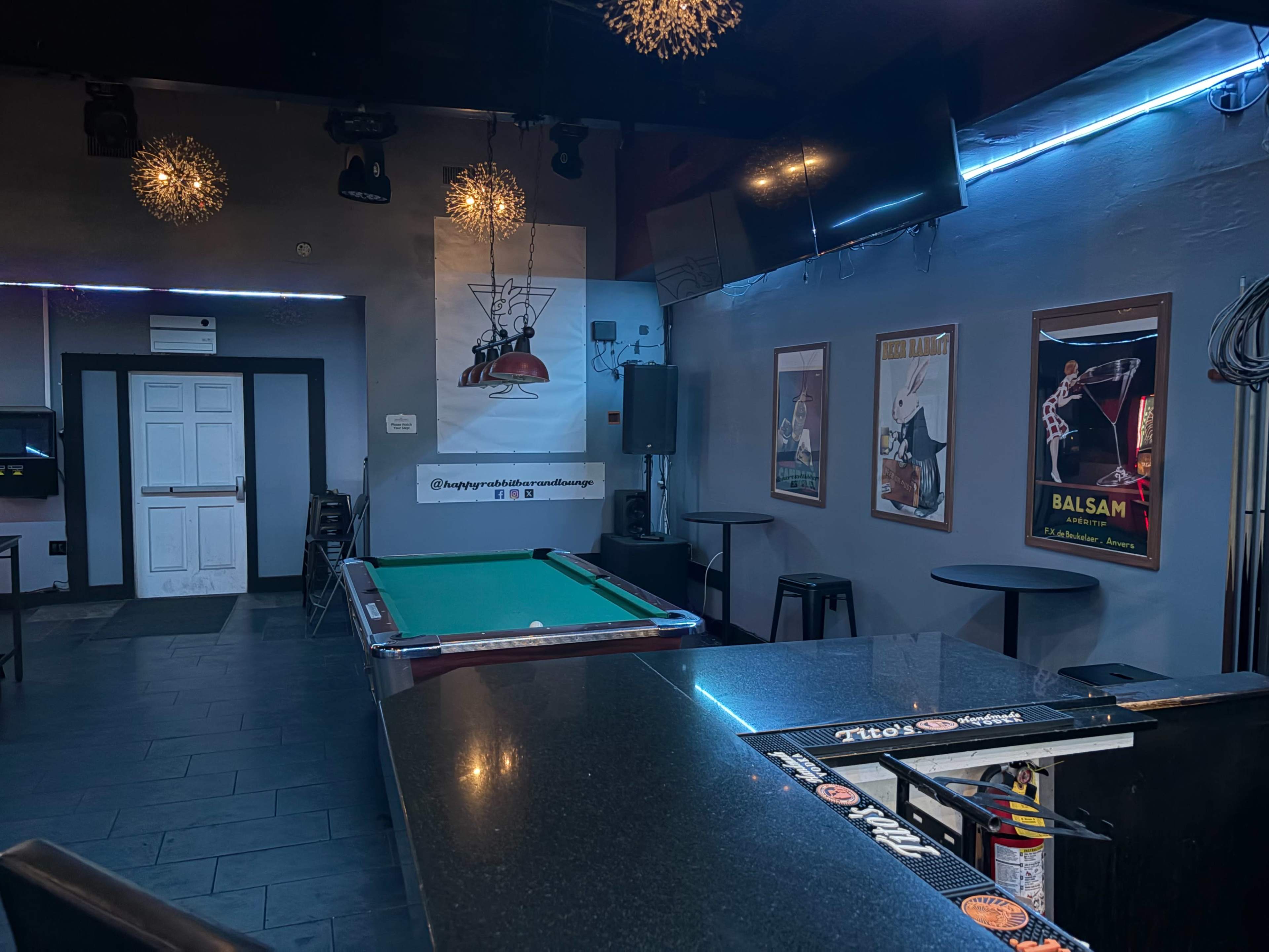 The image shows the interior of a bar with a pool table, a dark countertop, and several tables arranged around the area, along with framed posters on the walls.