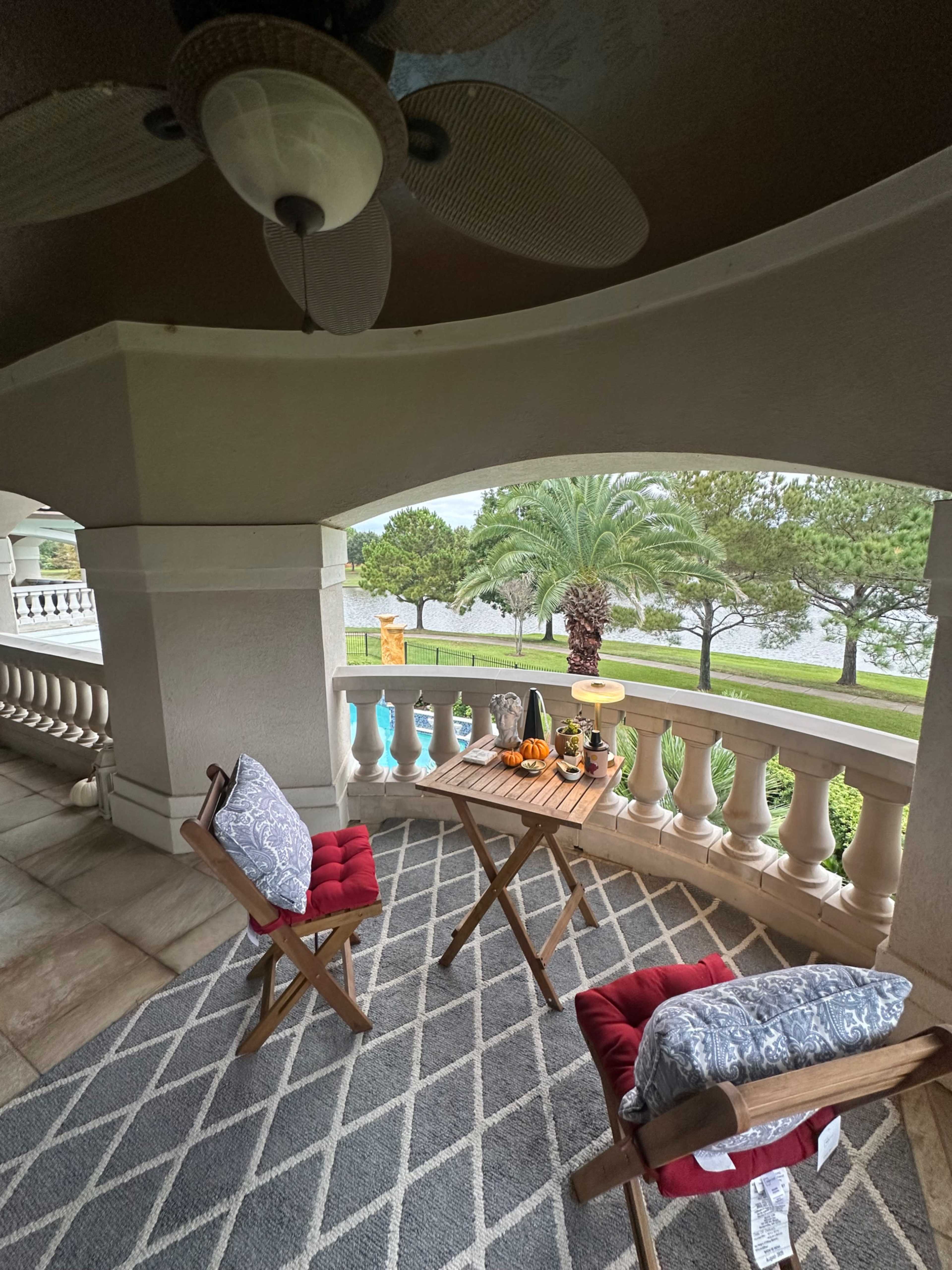 A cozy outdoor seating area features two red-cushioned chairs and a small wooden table, with a view of palm trees and a swimming pool beyond a decorative railing.