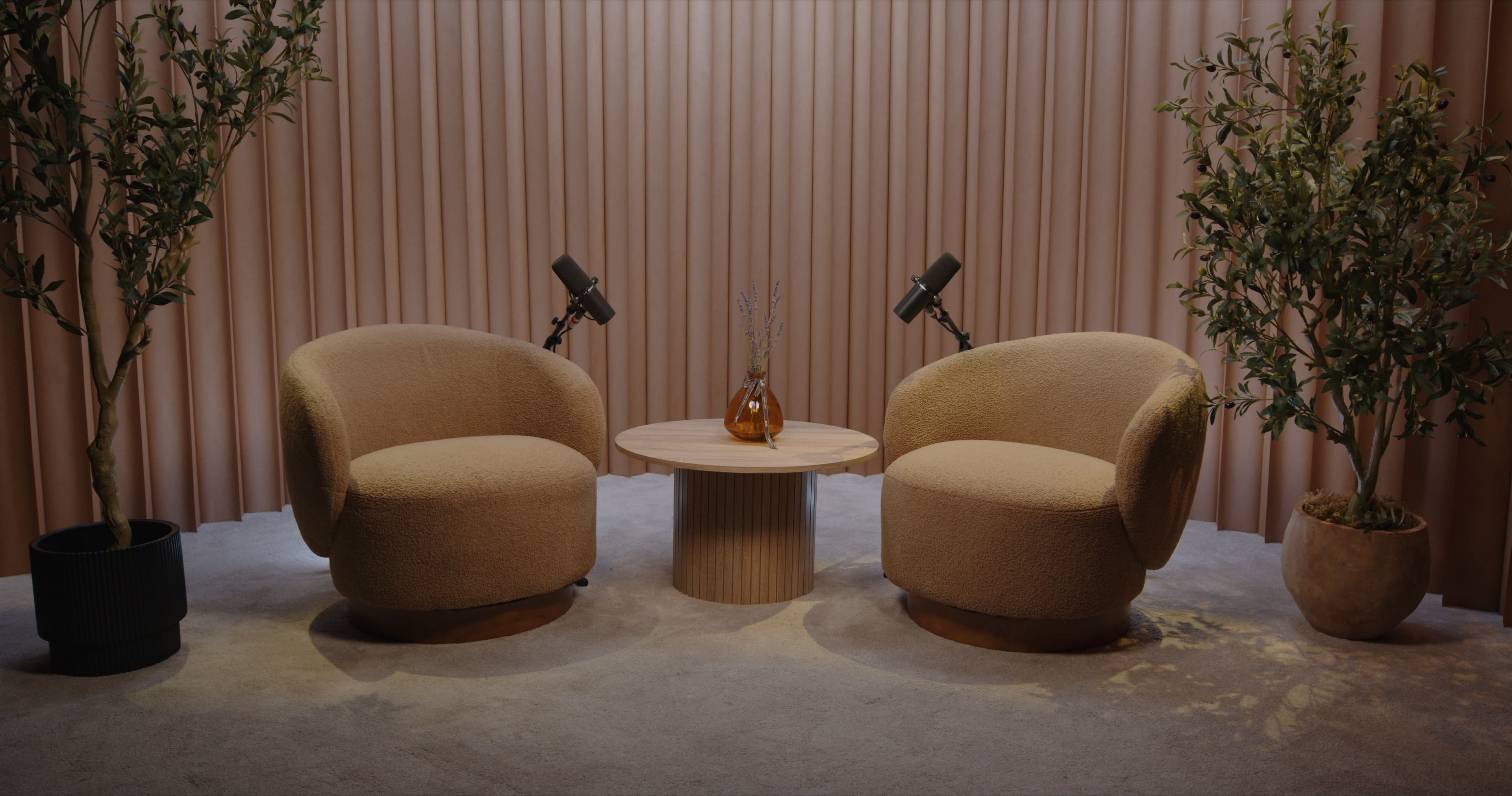 The image features a cozy seating arrangement with two beige armchairs facing each other, accompanied by a round wooden table and potted plants, all set against a backdrop of soft, vertical fabric panels.