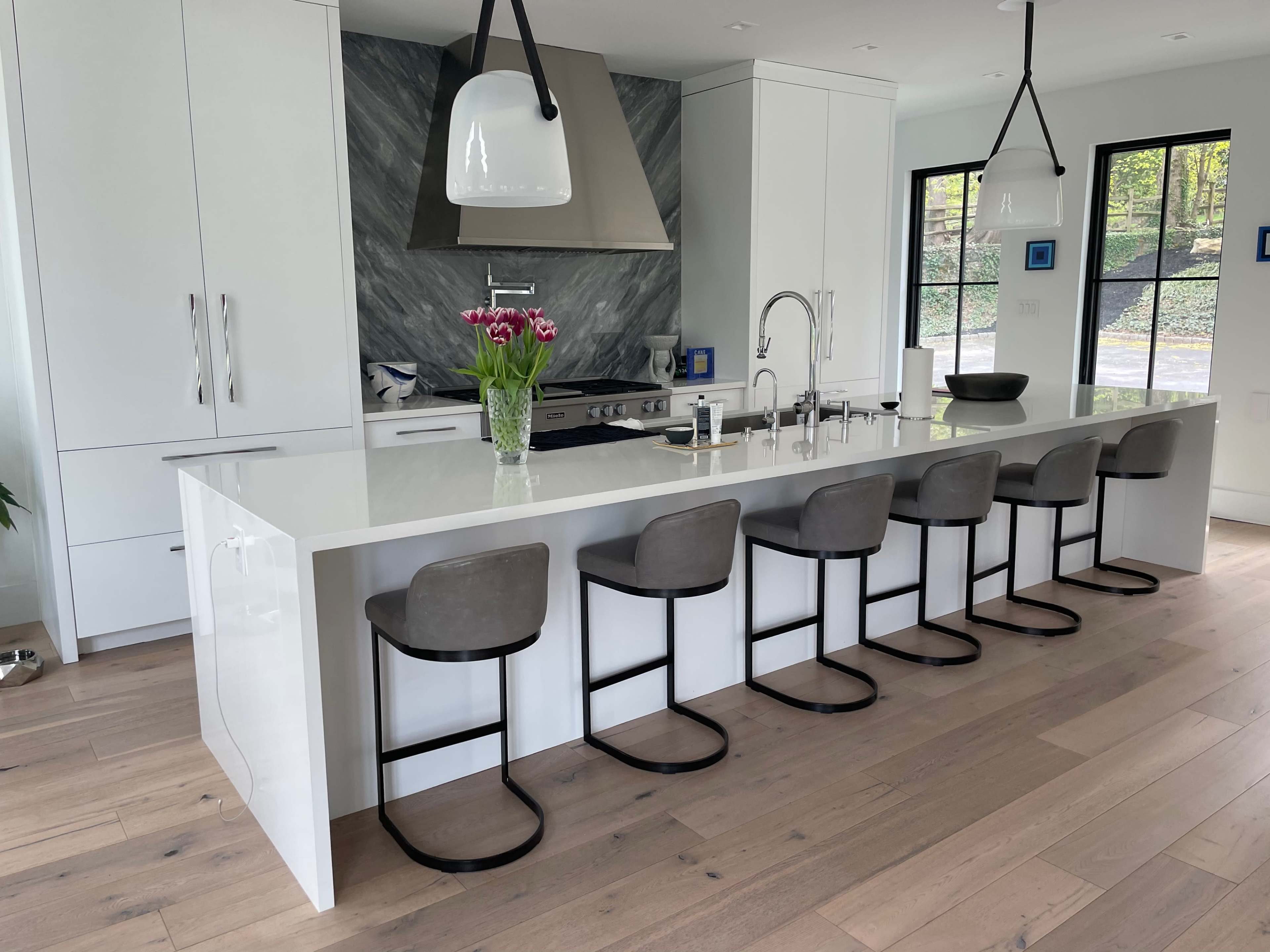 A modern kitchen with a white island countertop, six gray bar stools, and a marble backsplash behind a stainless steel stove.