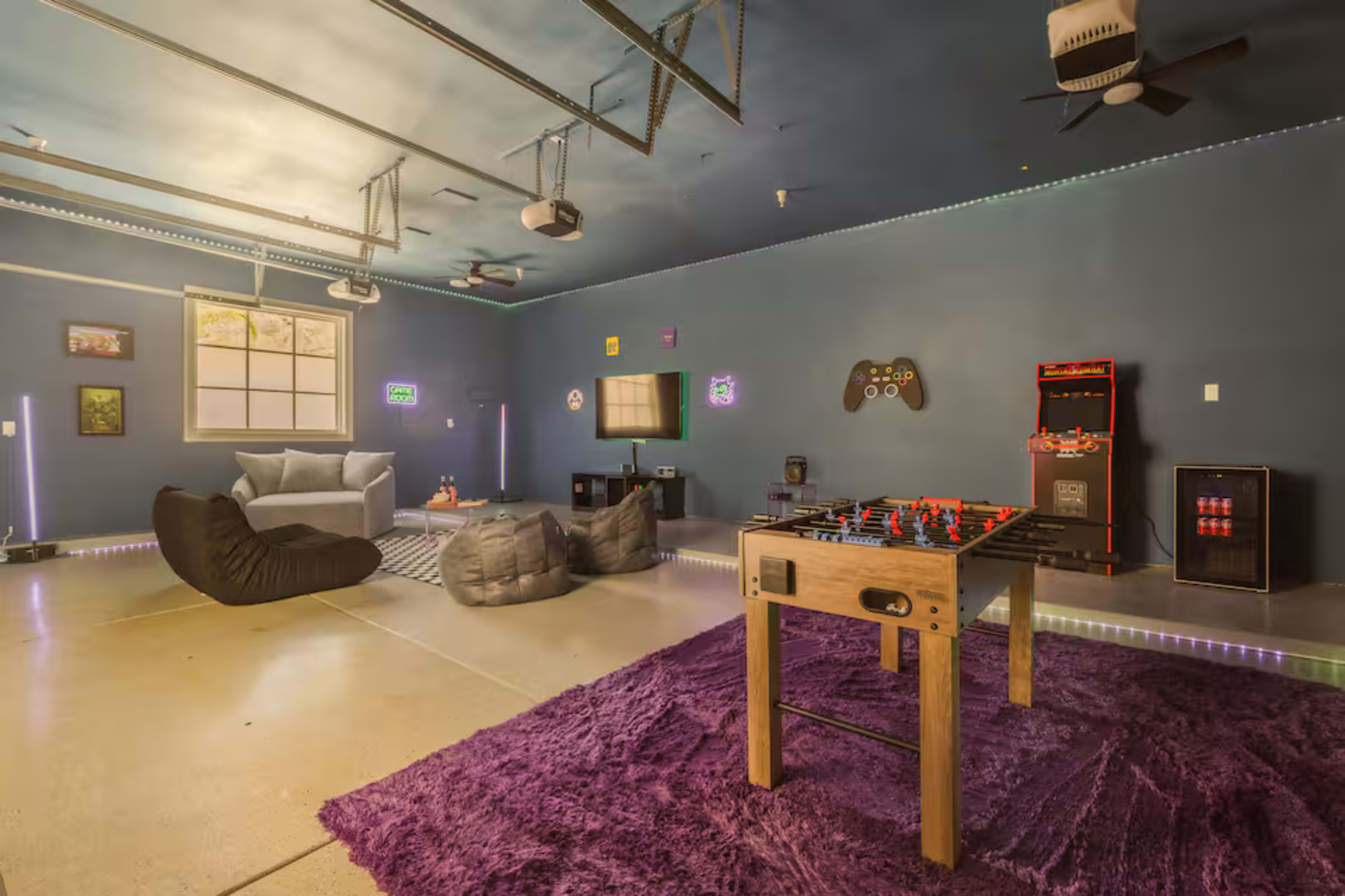 The image shows a spacious game room featuring a foosball table, comfortable seating, a television, and colorful wall decorations.