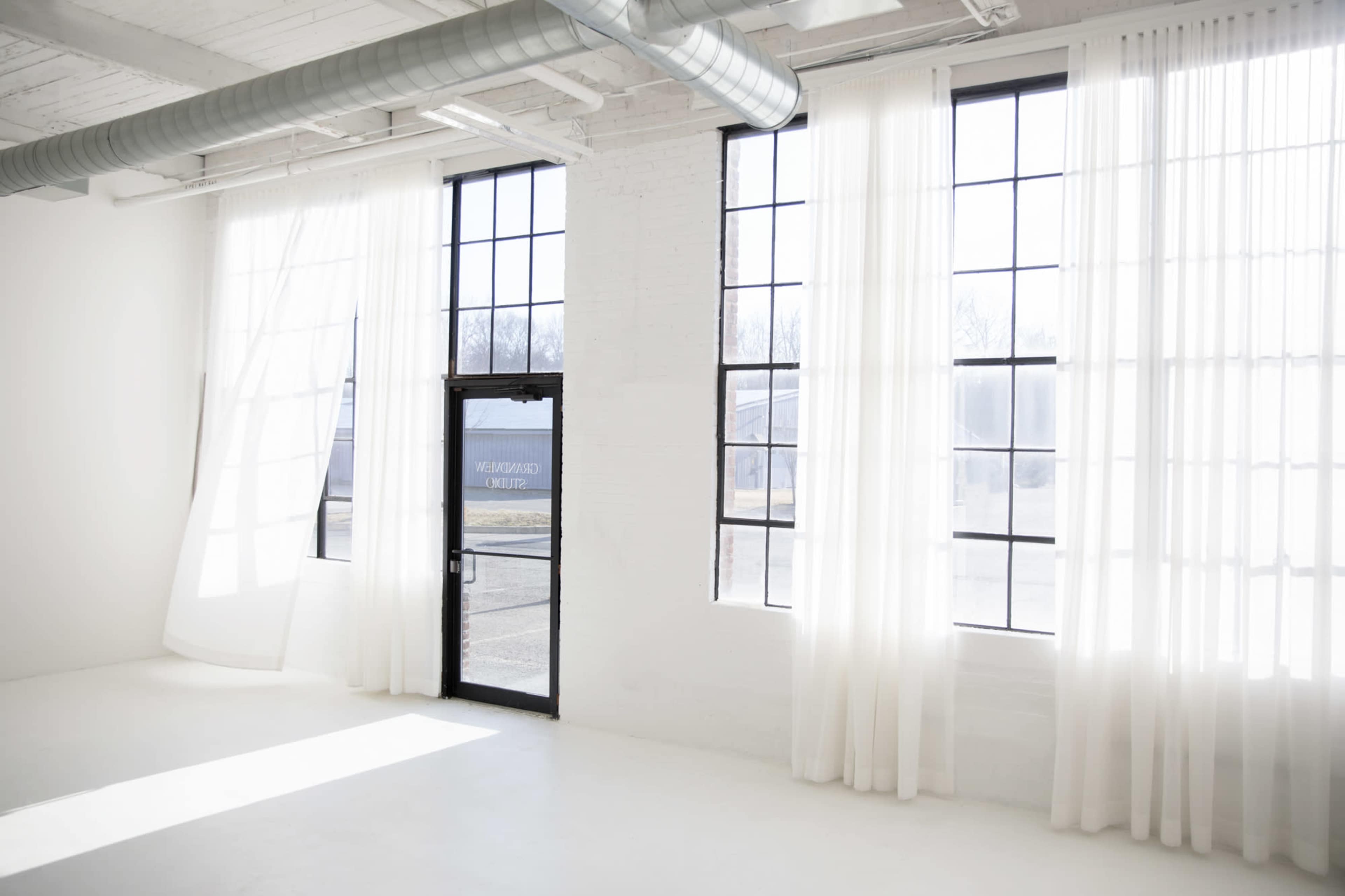 A sunlit interior features large windows framed by sheer curtains, with a door leading outside.
