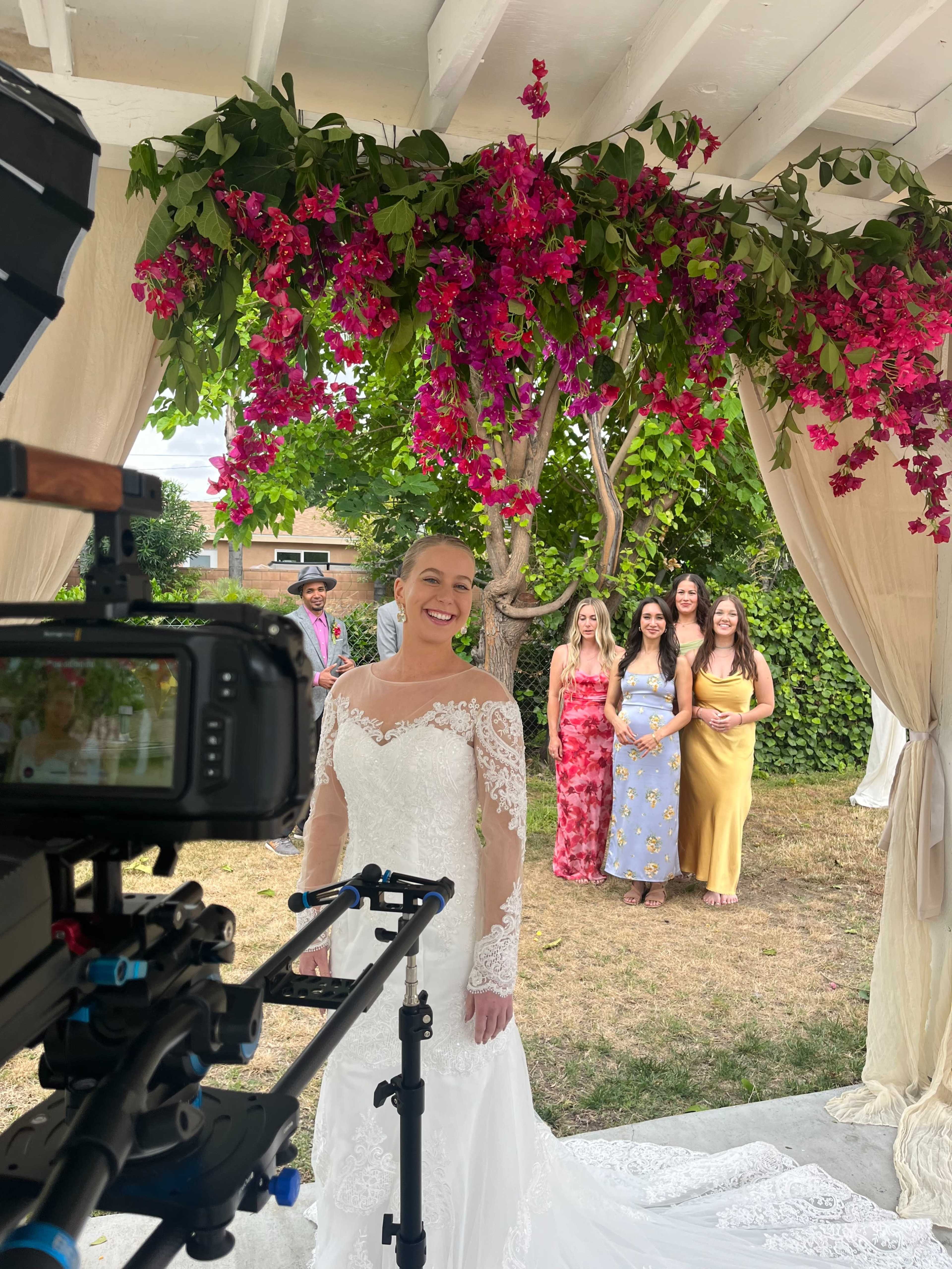 A bride in a white wedding dress smiles in front of a camera, with a backdrop of pink flowers and a group of women in colorful dresses standing behind her.
