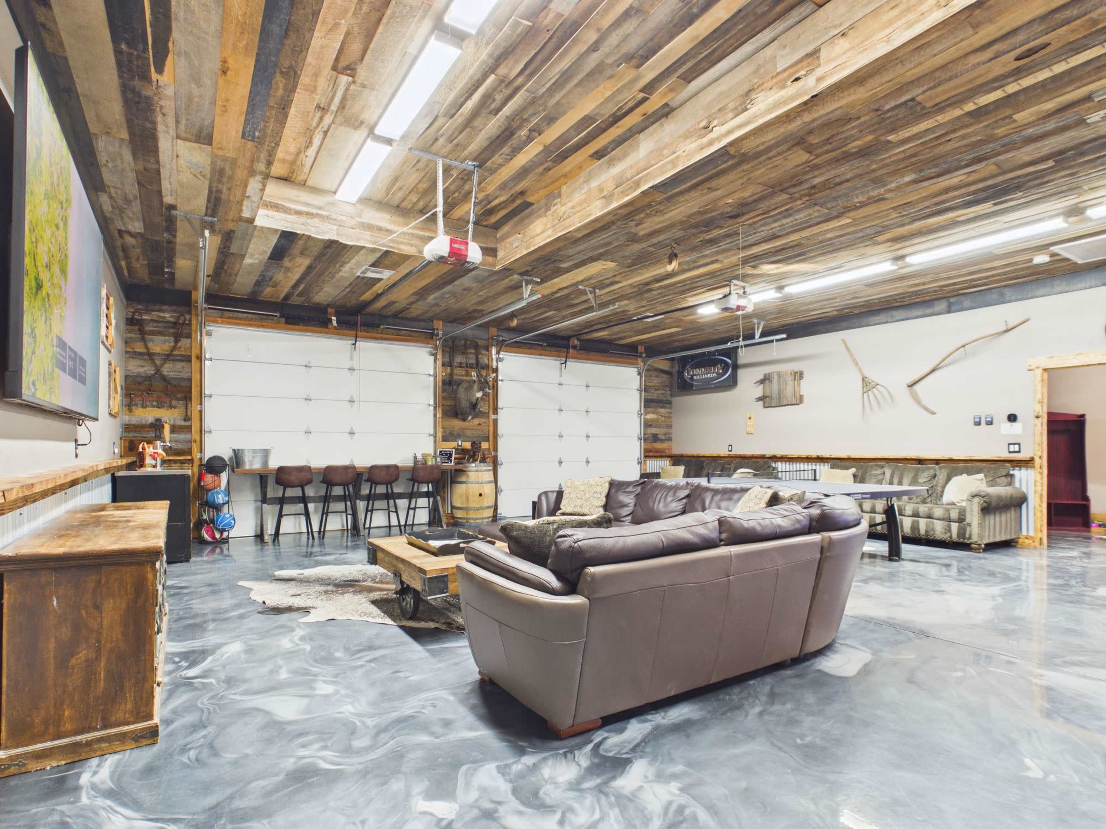 The image shows a spacious garage that has been converted into a comfortable lounge area with dark leather sofas, a concrete floor, and wooden ceiling beams.