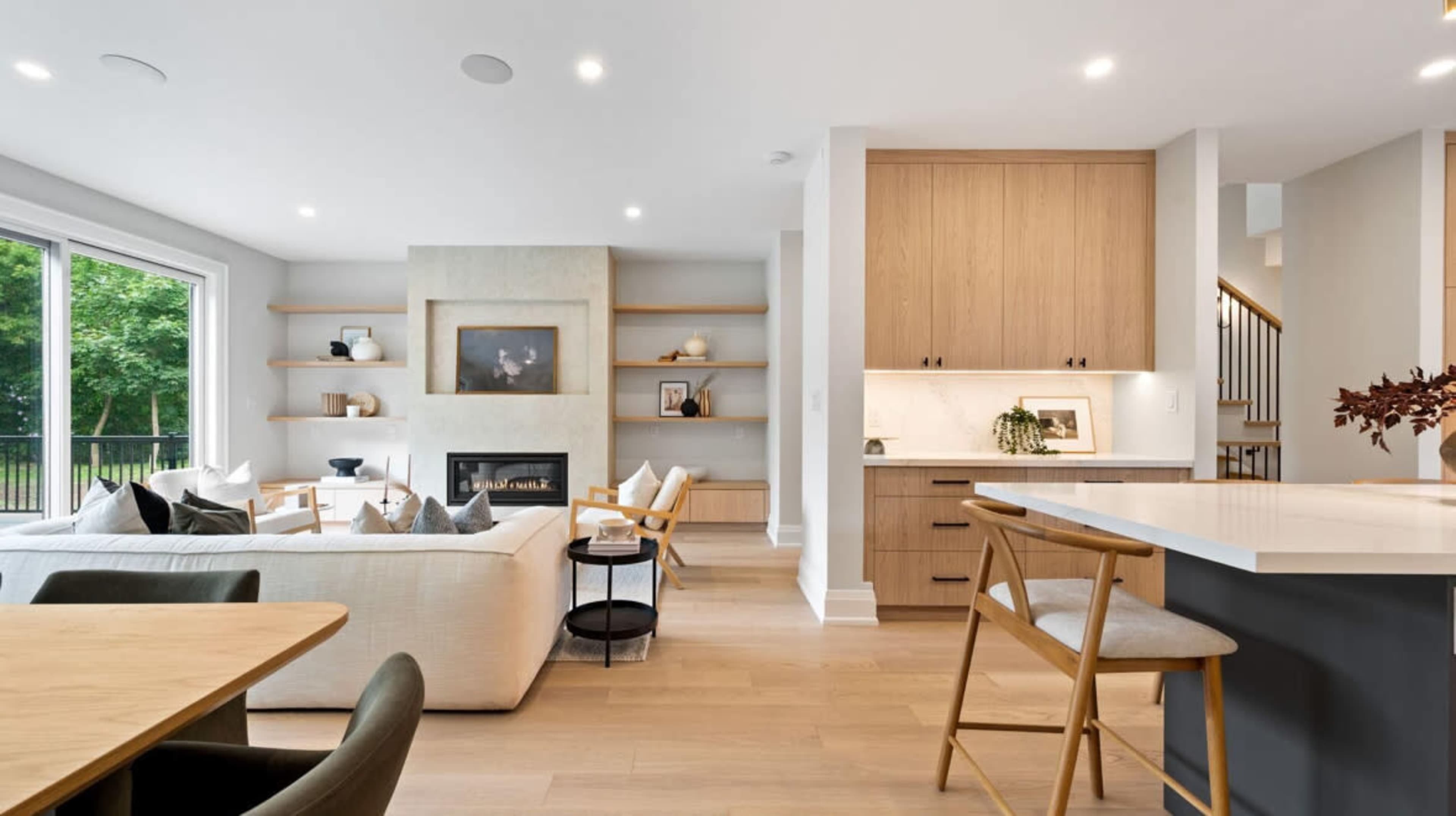 The image shows a modern living room with a light color palette, featuring a fireplace, shelving, and an open kitchen with wooden cabinets and a dining area.