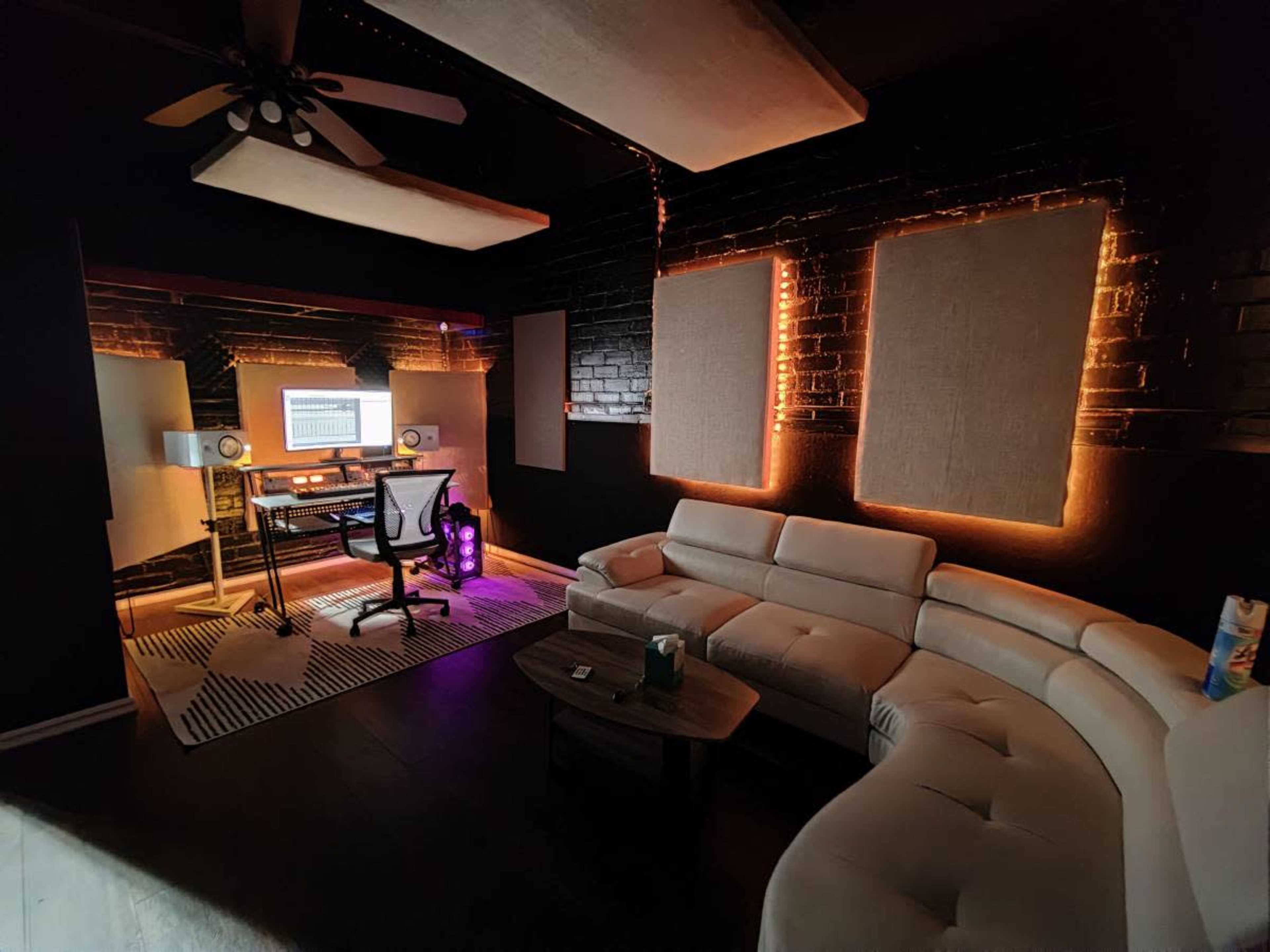 The image shows a modern recording studio with a curved white sofa, a wooden table, and a music workstation featuring a computer and studio monitors.