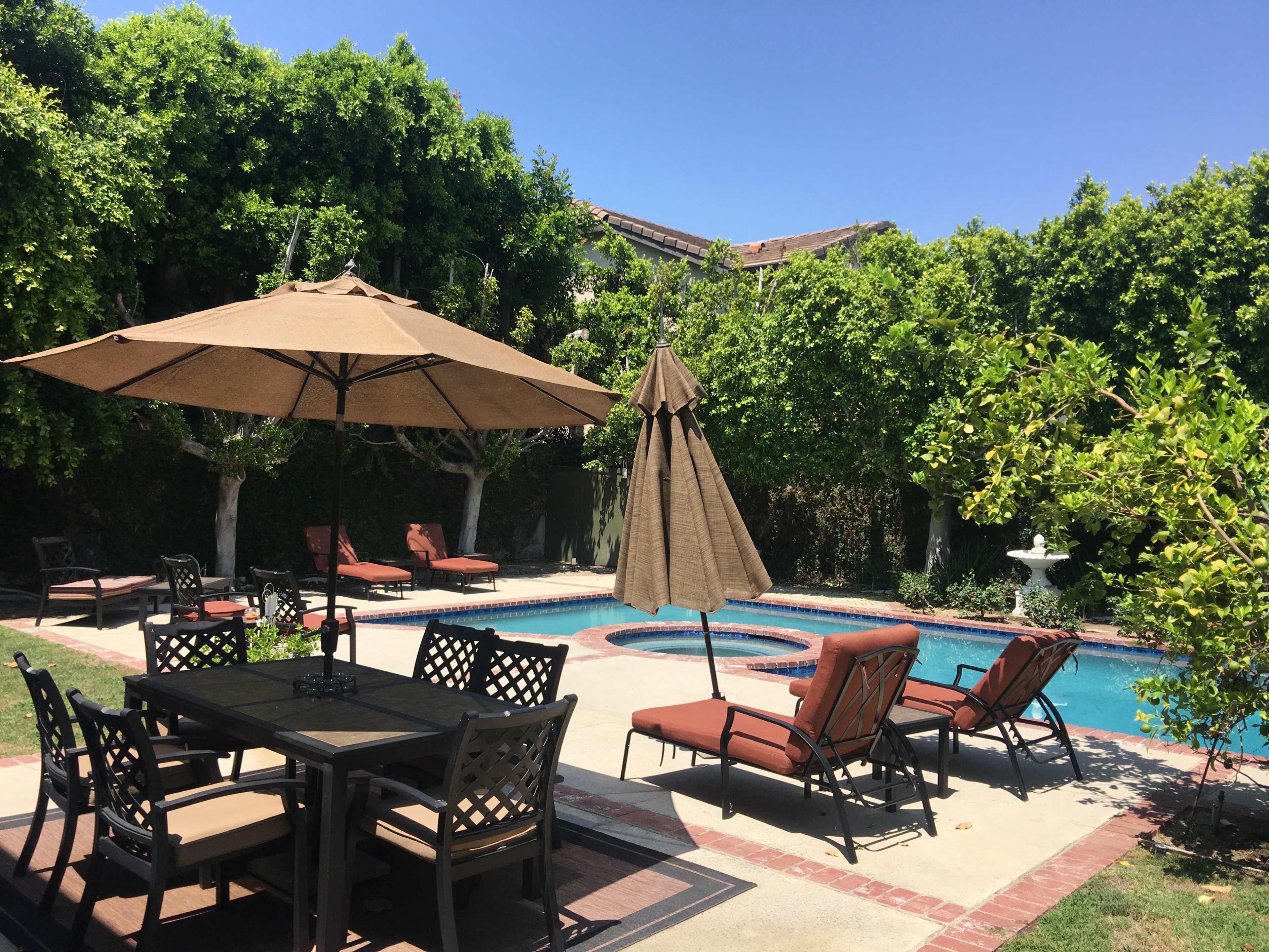 The image shows a backyard pool area with lounge chairs, a dining table, and umbrellas surrounded by green foliage.