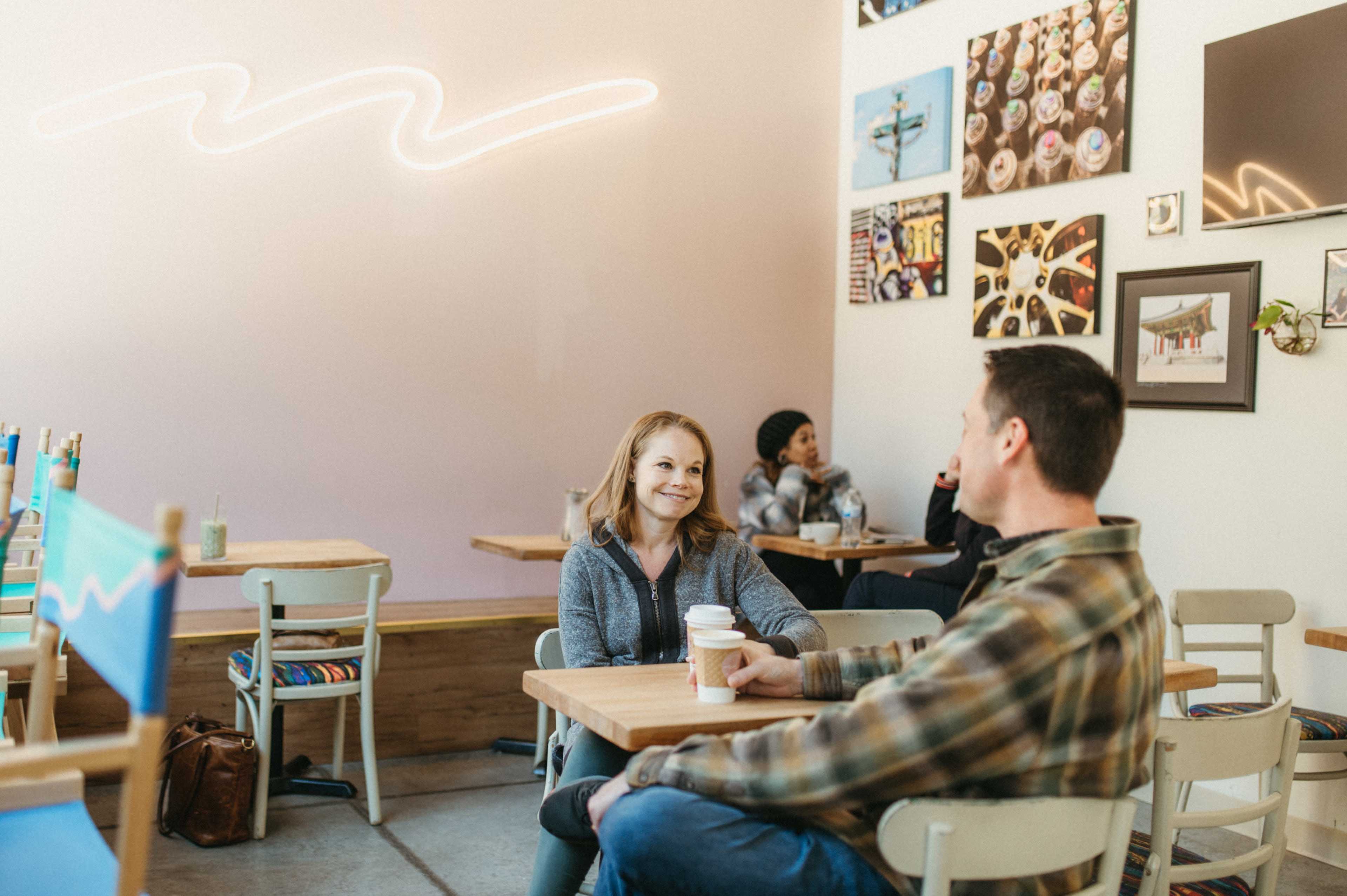Two people sit at a table in a cafe, engaging in conversation, while other customers are seen in the background.