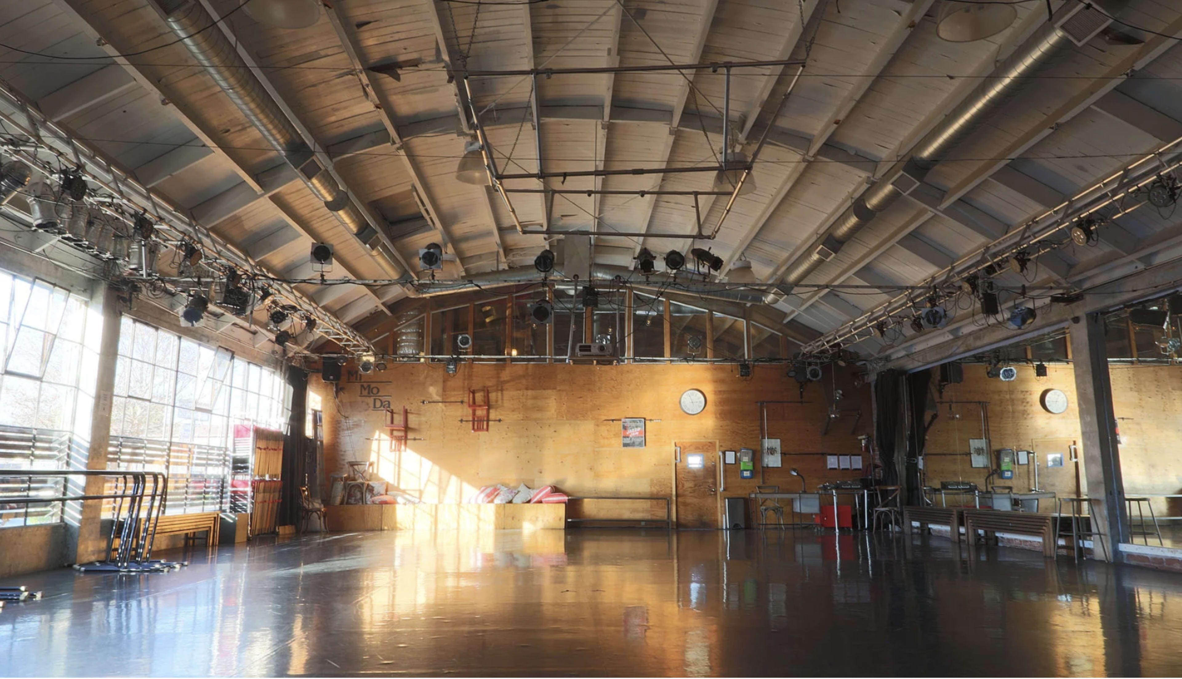 The image shows an empty gymnasium with a wooden floor, large windows, and overhead lighting.