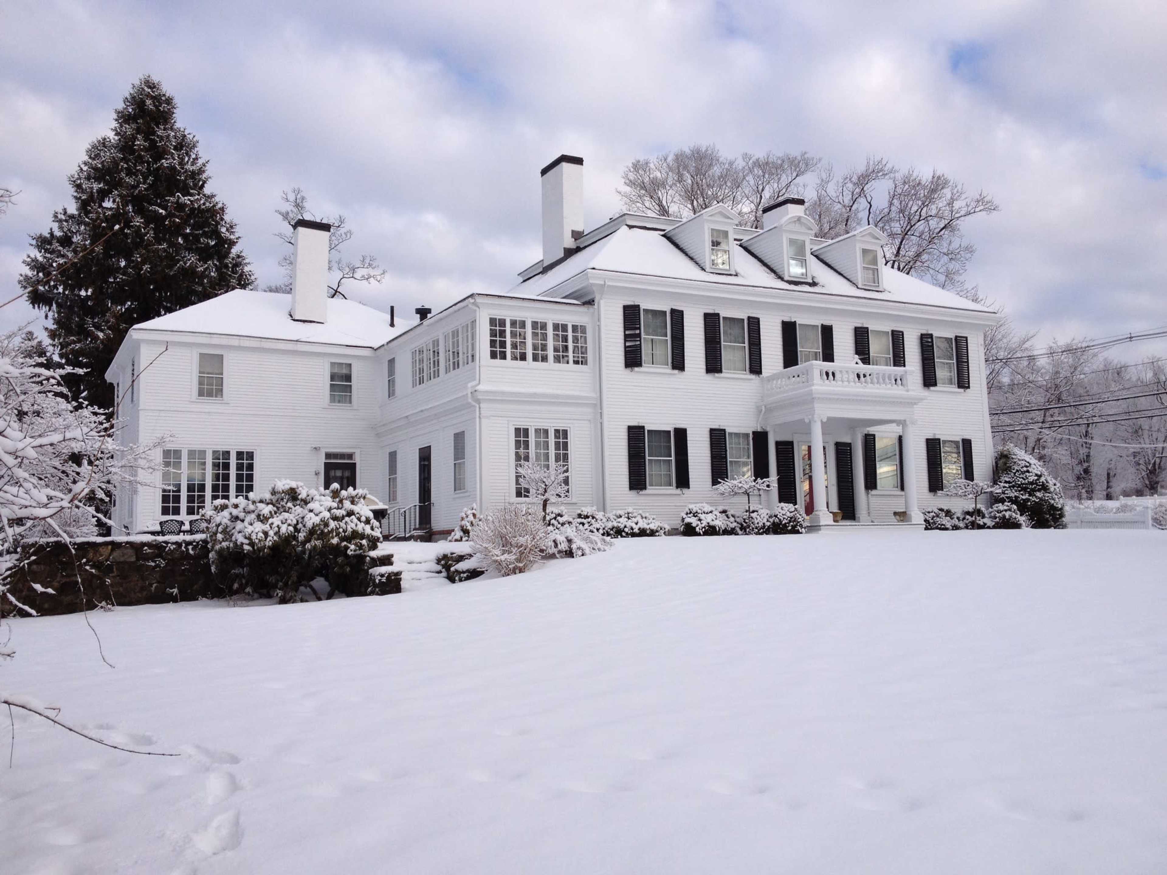 A large white colonial-style house sits surrounded by freshly fallen snow under a cloudy sky.