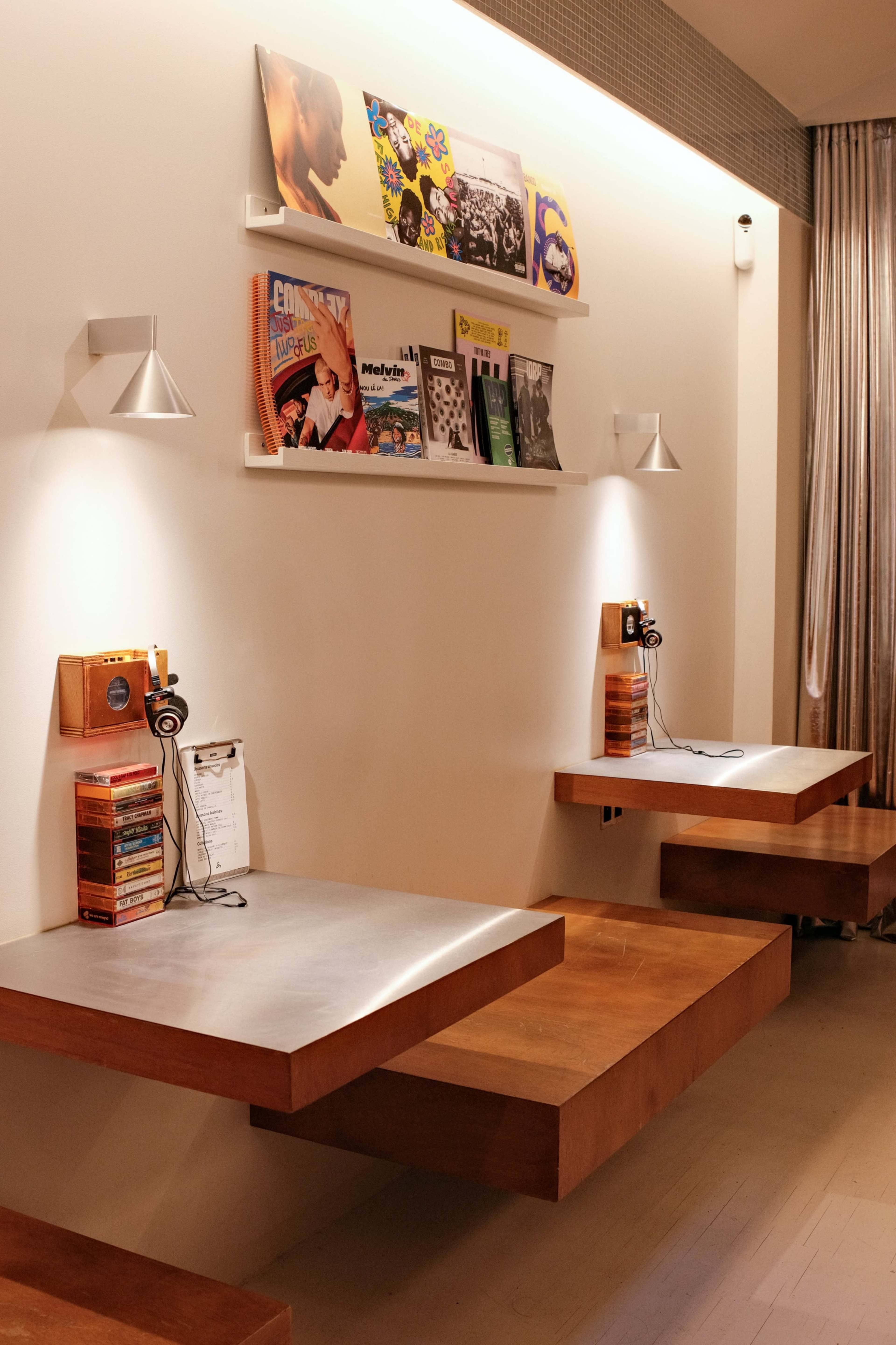 The image shows a minimalist interior with wooden tables attached to a white wall, displaying vinyl records and providing headphone connections for listening.