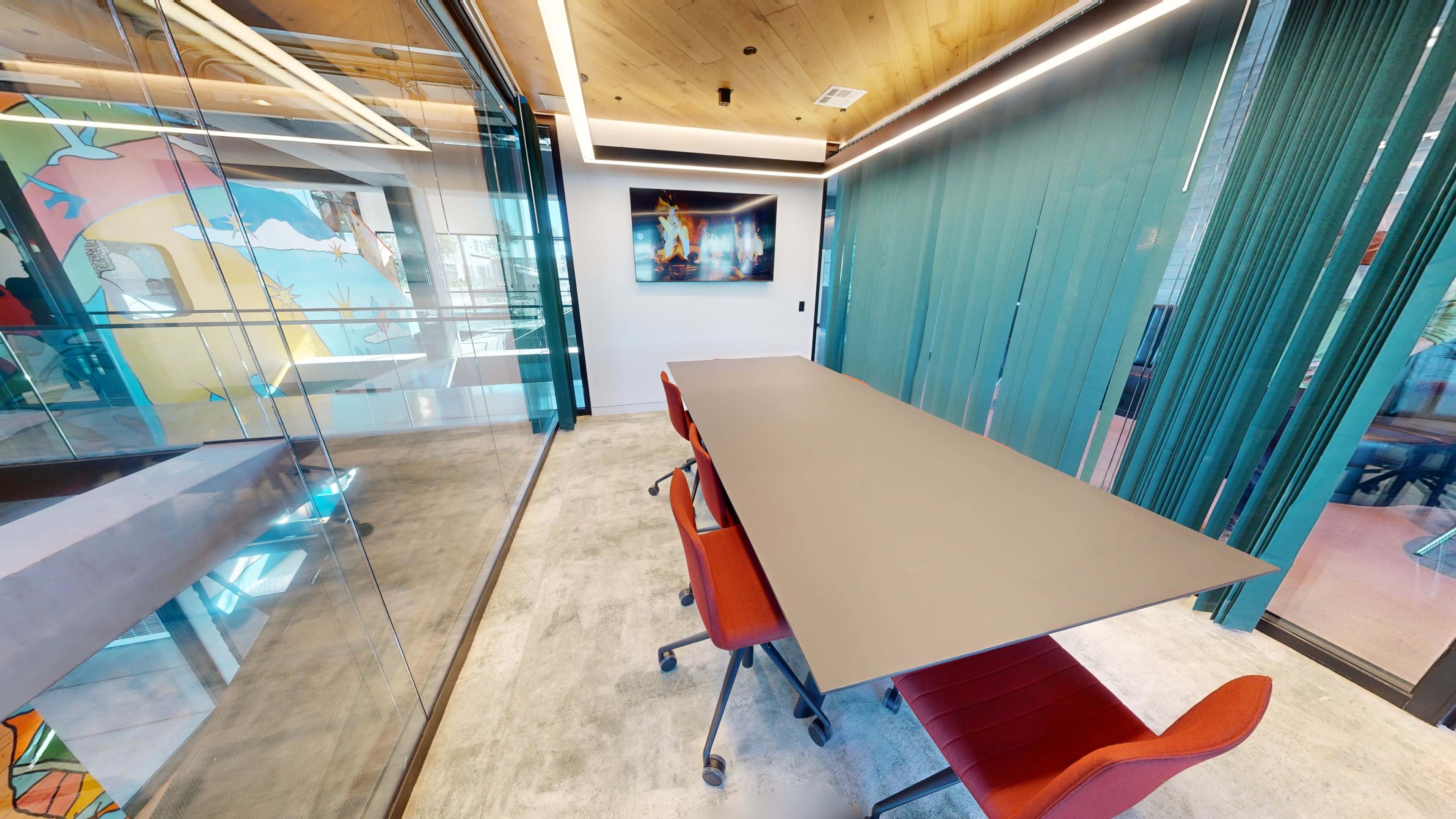 A modern conference room features a long table flanked by red chairs, with glass walls and colorful artwork in the background.