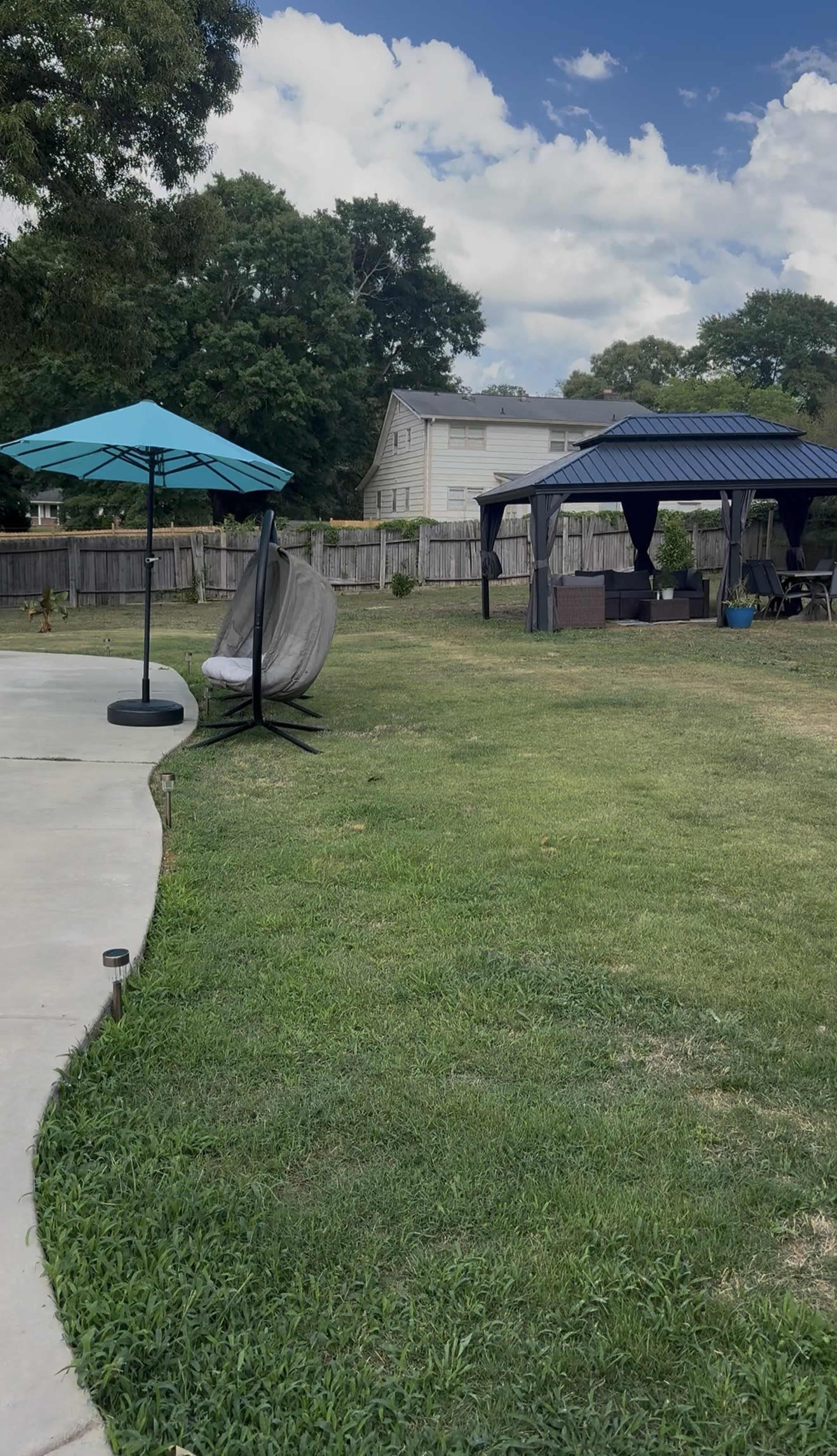 The image shows a backyard with a curved concrete path, a turquoise patio umbrella, a hanging chair, and two shaded seating areas under canopies.