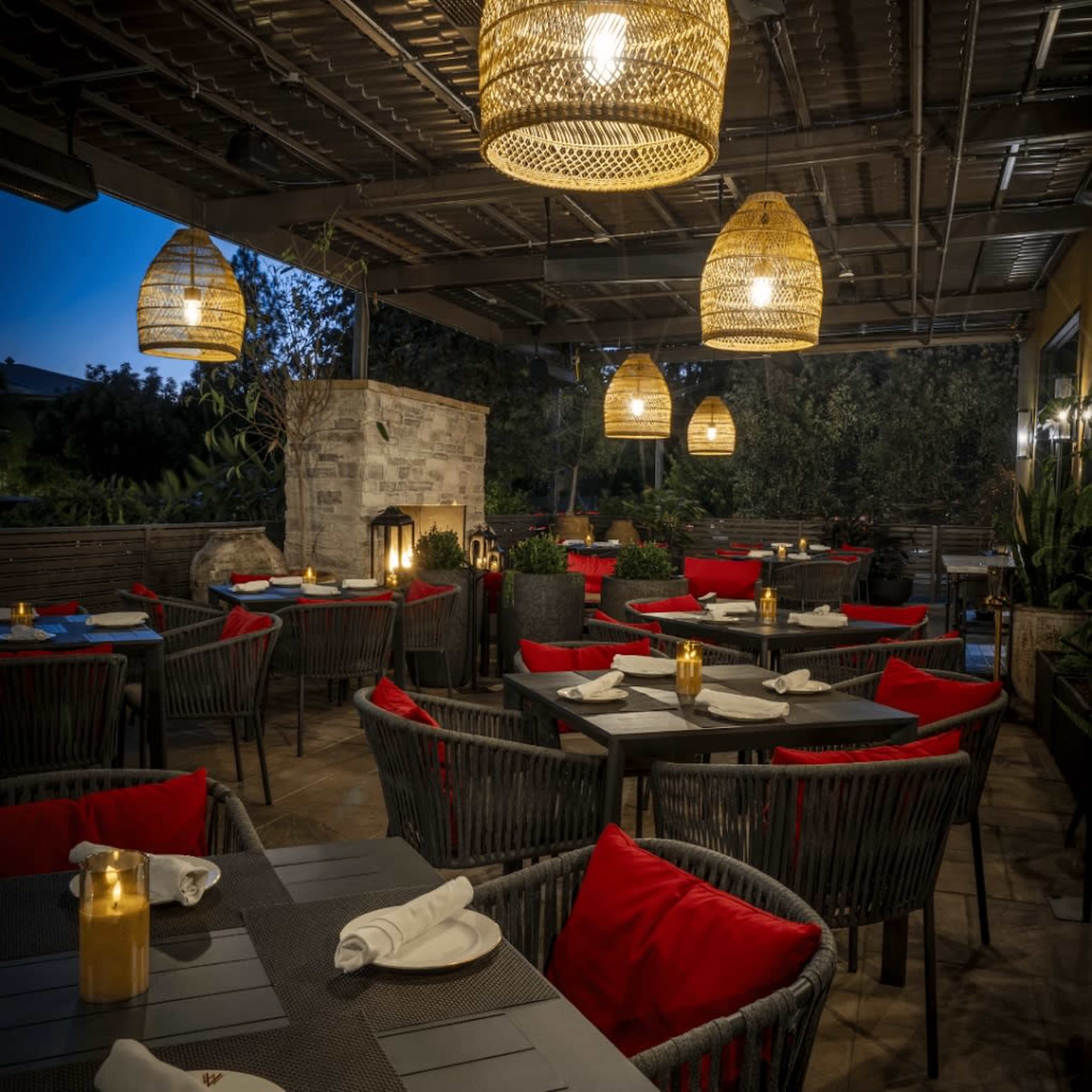 The image shows an outdoor restaurant patio with wicker furniture, red cushions, and hanging lights against a twilight background.