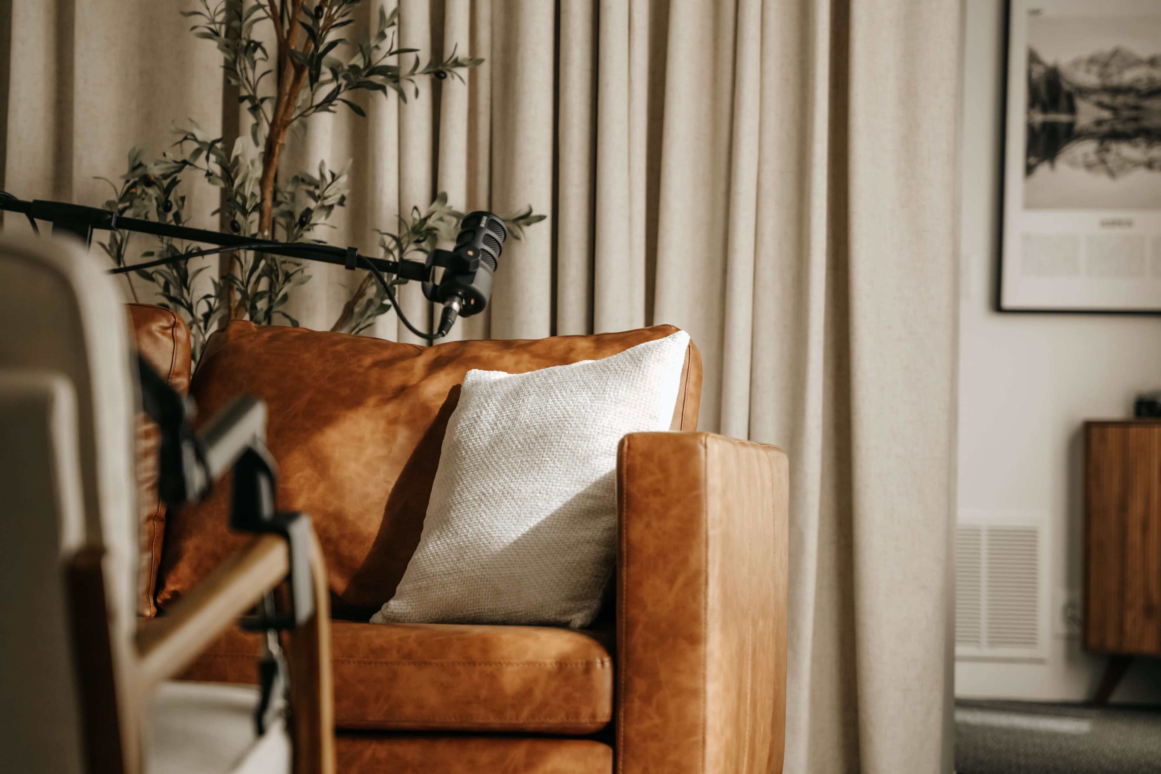 A brown leather couch with a white textured pillow is positioned near a curtain and a floor lamp in a cozy living room setting.