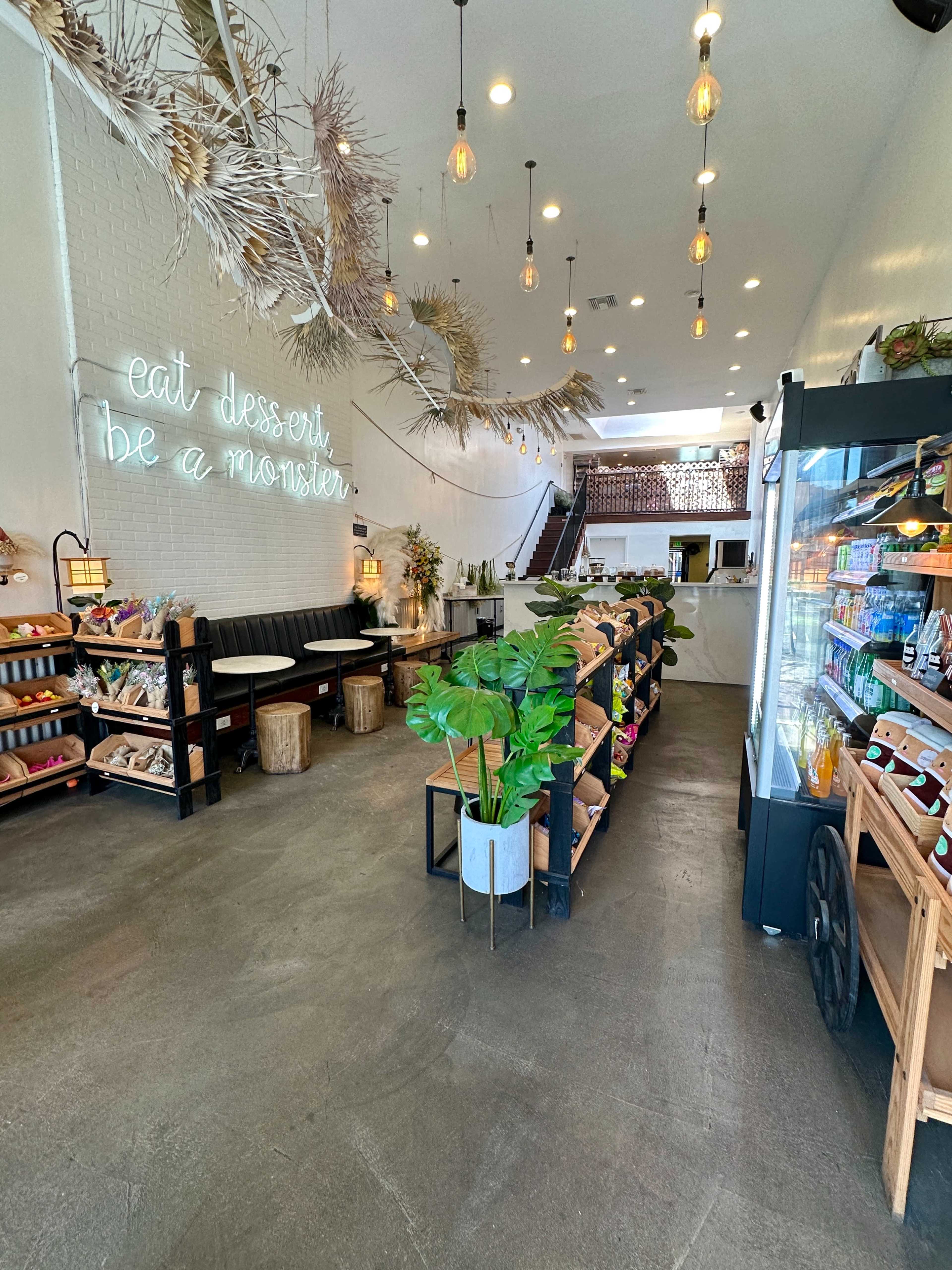 The image shows a brightly lit dessert shop with a minimalist design, featuring shelves filled with various desserts and a neon sign that reads "eat dessert be a monster."