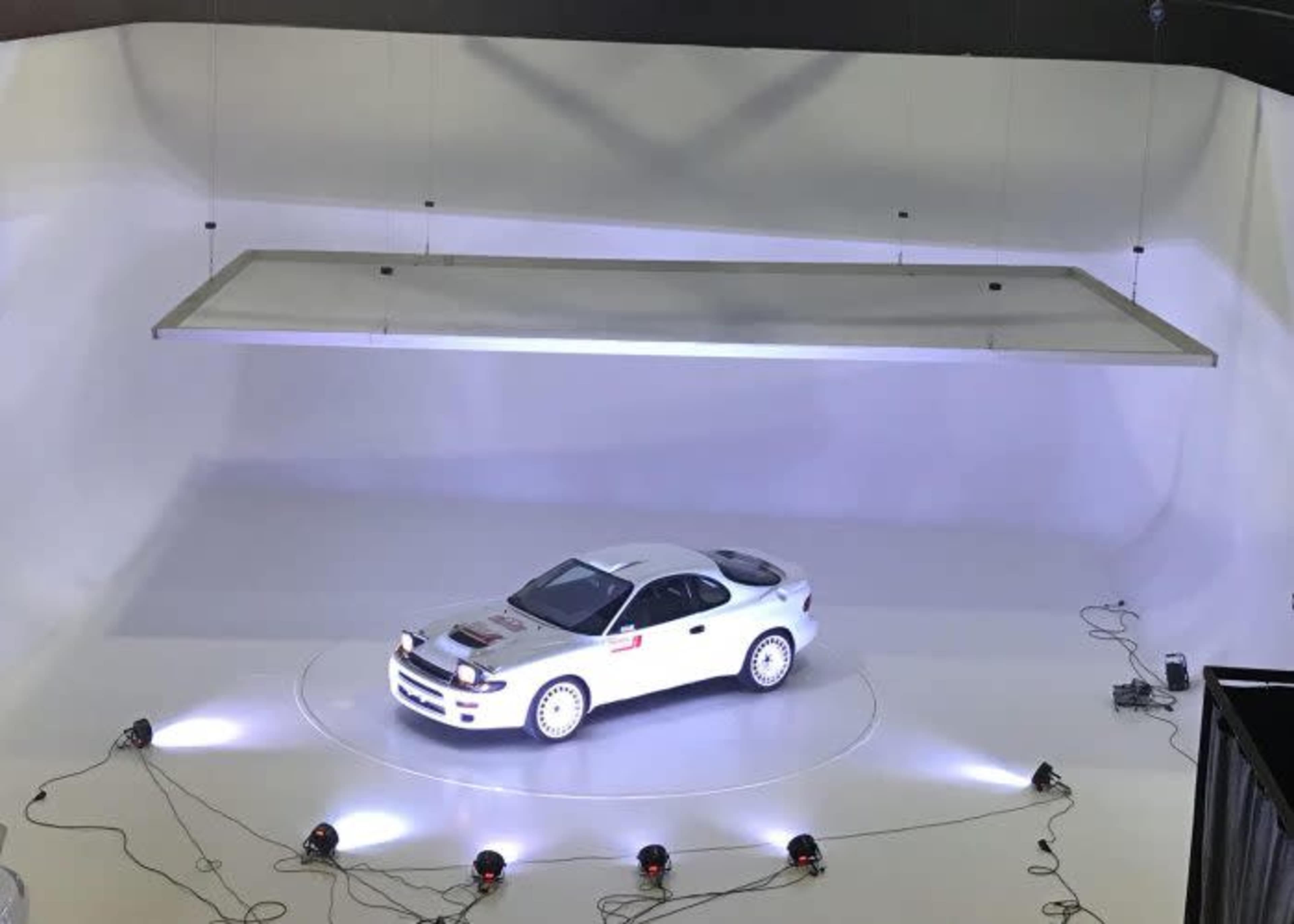 A white sports car is shown on a circular platform under bright lights in a minimalistic studio setting.