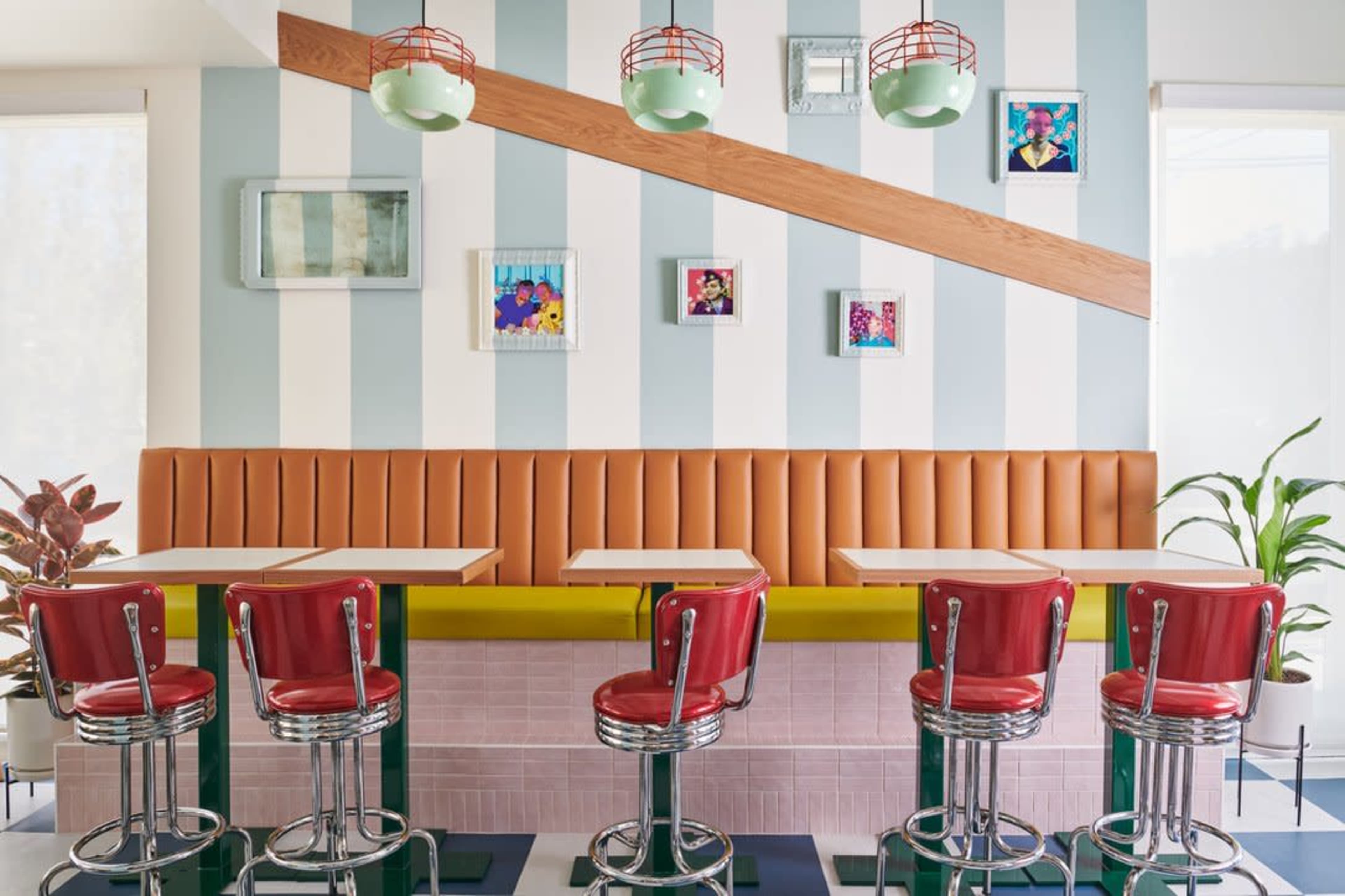 A retro-style diner interior with red bar stools lined up against a green and orange banquette, featuring striped walls and colorful artwork.