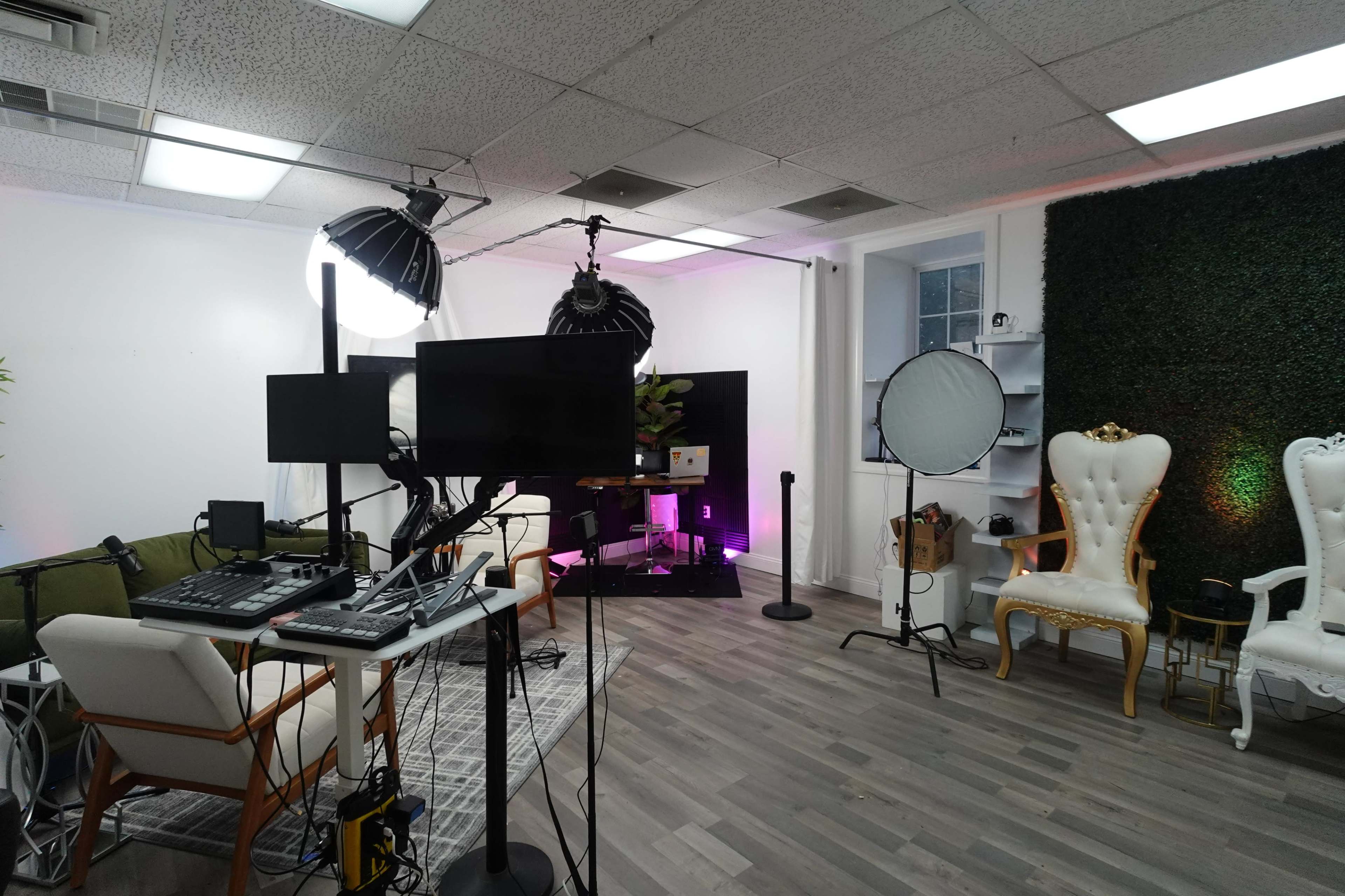 A modern studio space with green plants, multiple screens, lighting equipment, and seating arranged for filming or photography.