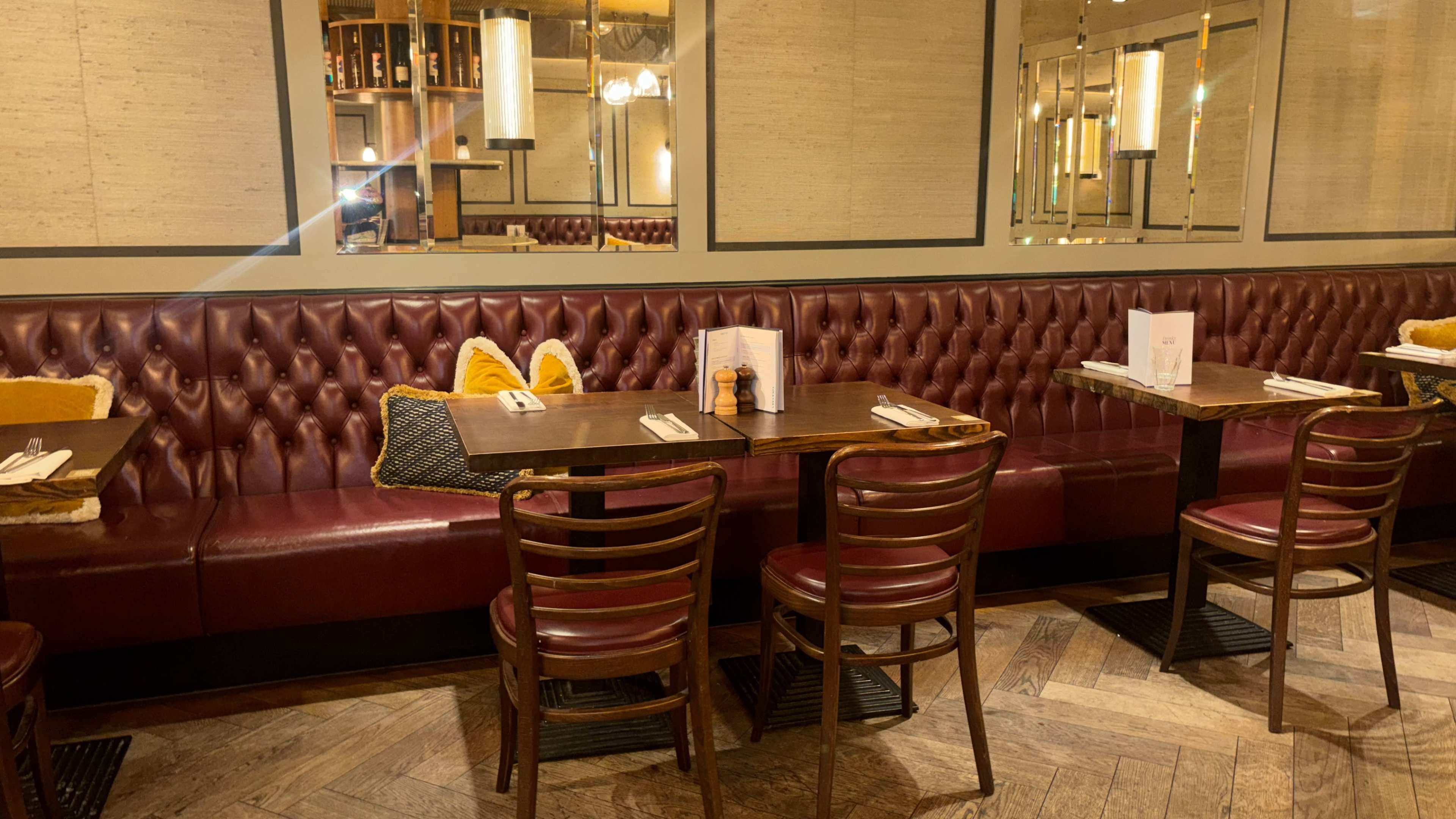 The image shows a cozy restaurant interior featuring maroon upholstered seating, wooden tables, and mirrors on the walls.