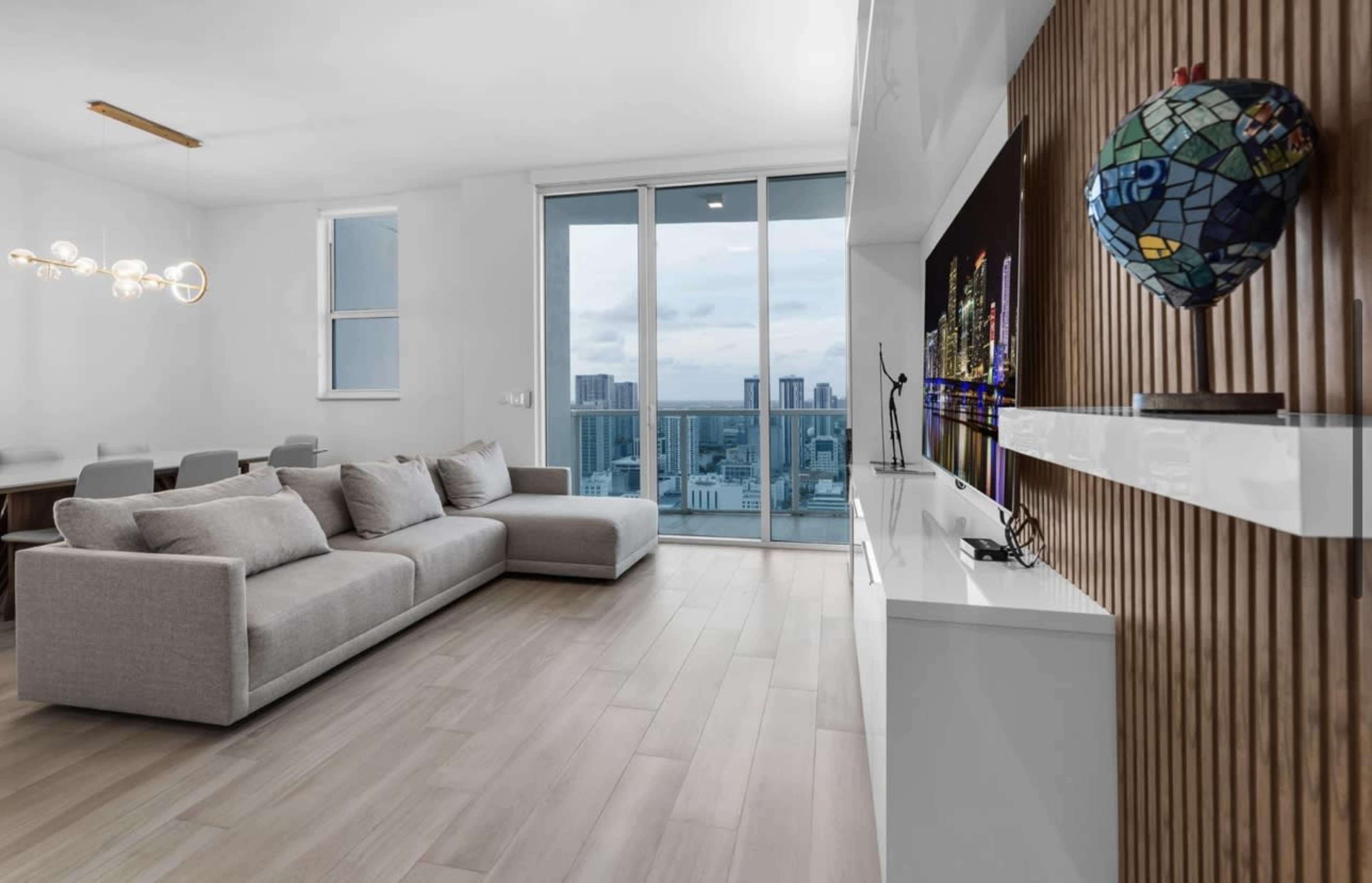 A modern living room with a large gray sectional sofa, a white entertainment unit, and a view of the city skyline through floor-to-ceiling windows.