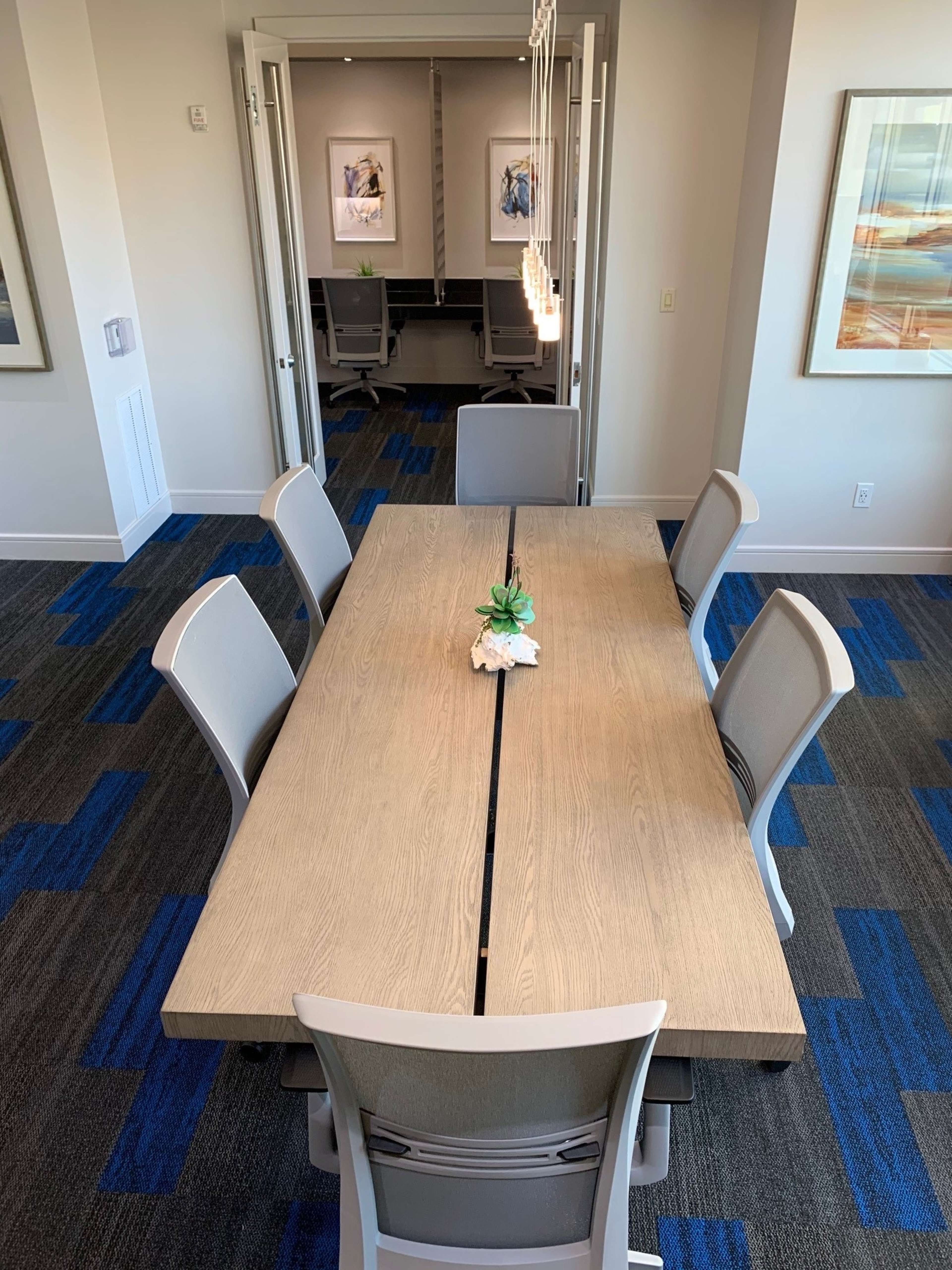 A long wooden table with six chairs is centered in a brightly lit conference room, featuring a small plant centerpiece and modern decor.