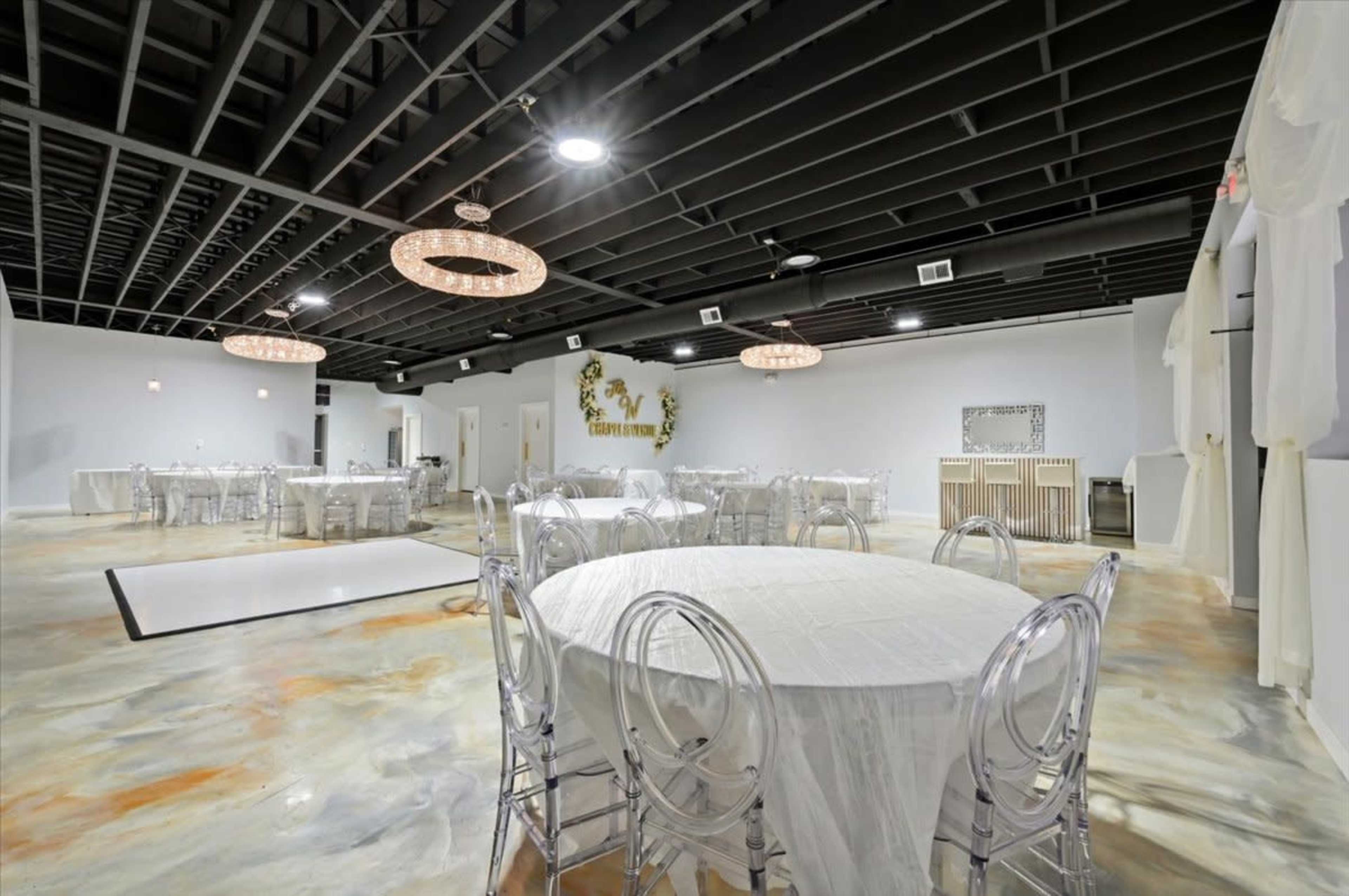 The image shows a spacious, well-lit event venue with round tables covered in white tablecloths and transparent chairs arranged throughout the room.