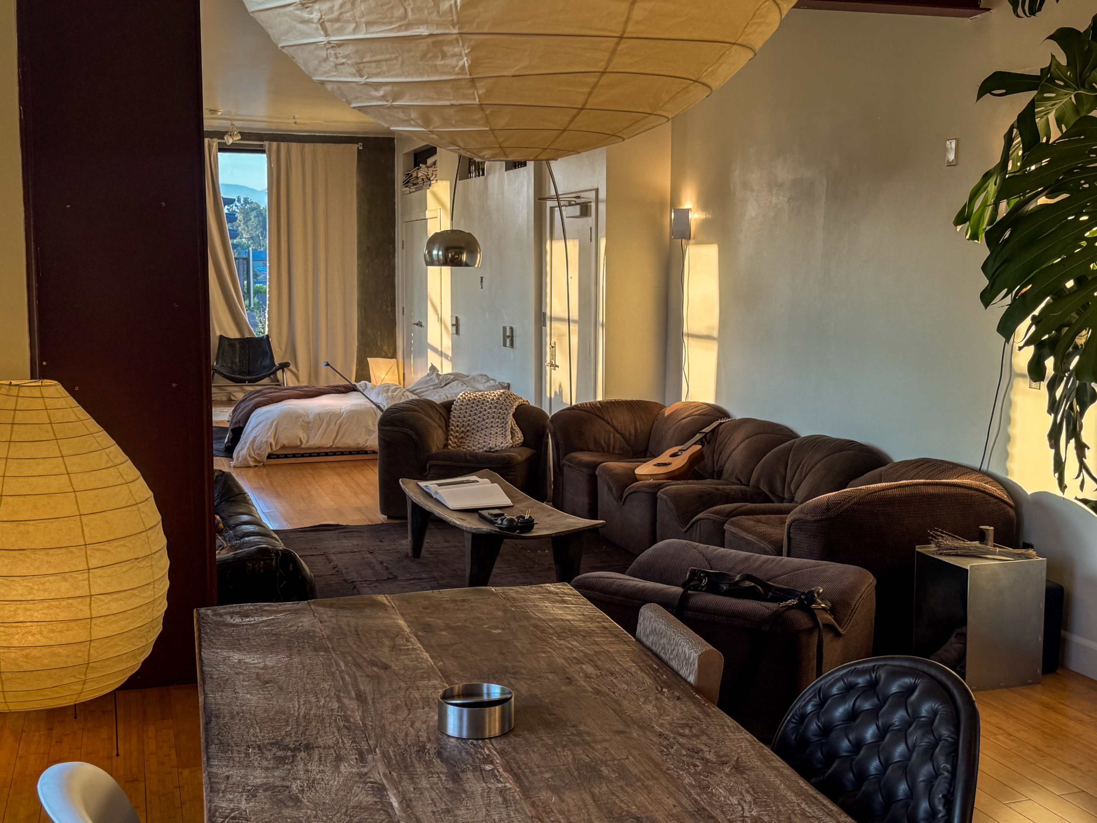 The image shows a spacious, modern living area with a seating arrangement of dark couches, a wooden dining table, and large windows allowing natural light to fill the space.