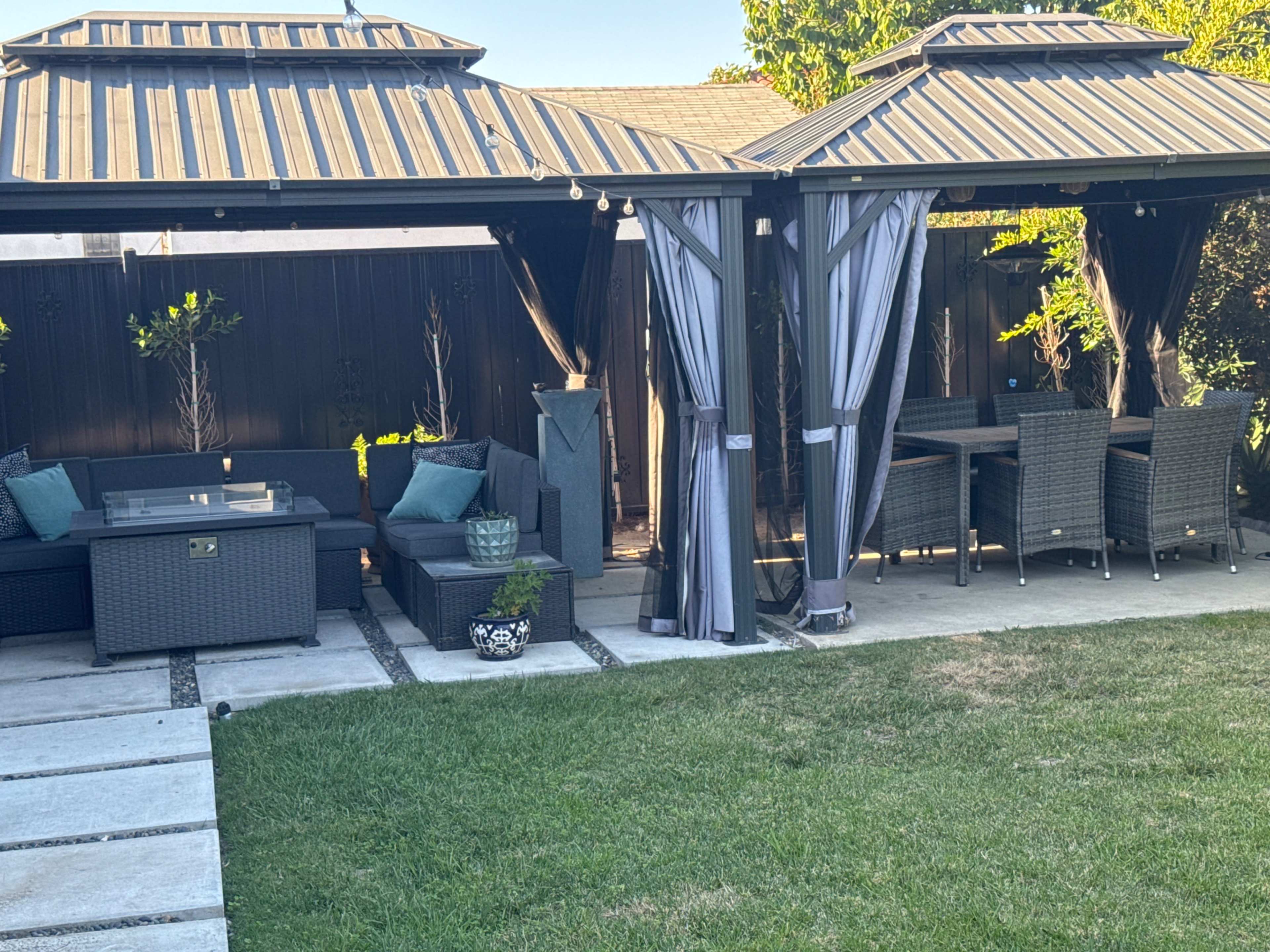 A backyard features a covered seating area with dark wicker furniture, a dining table, and a neatly maintained lawn.