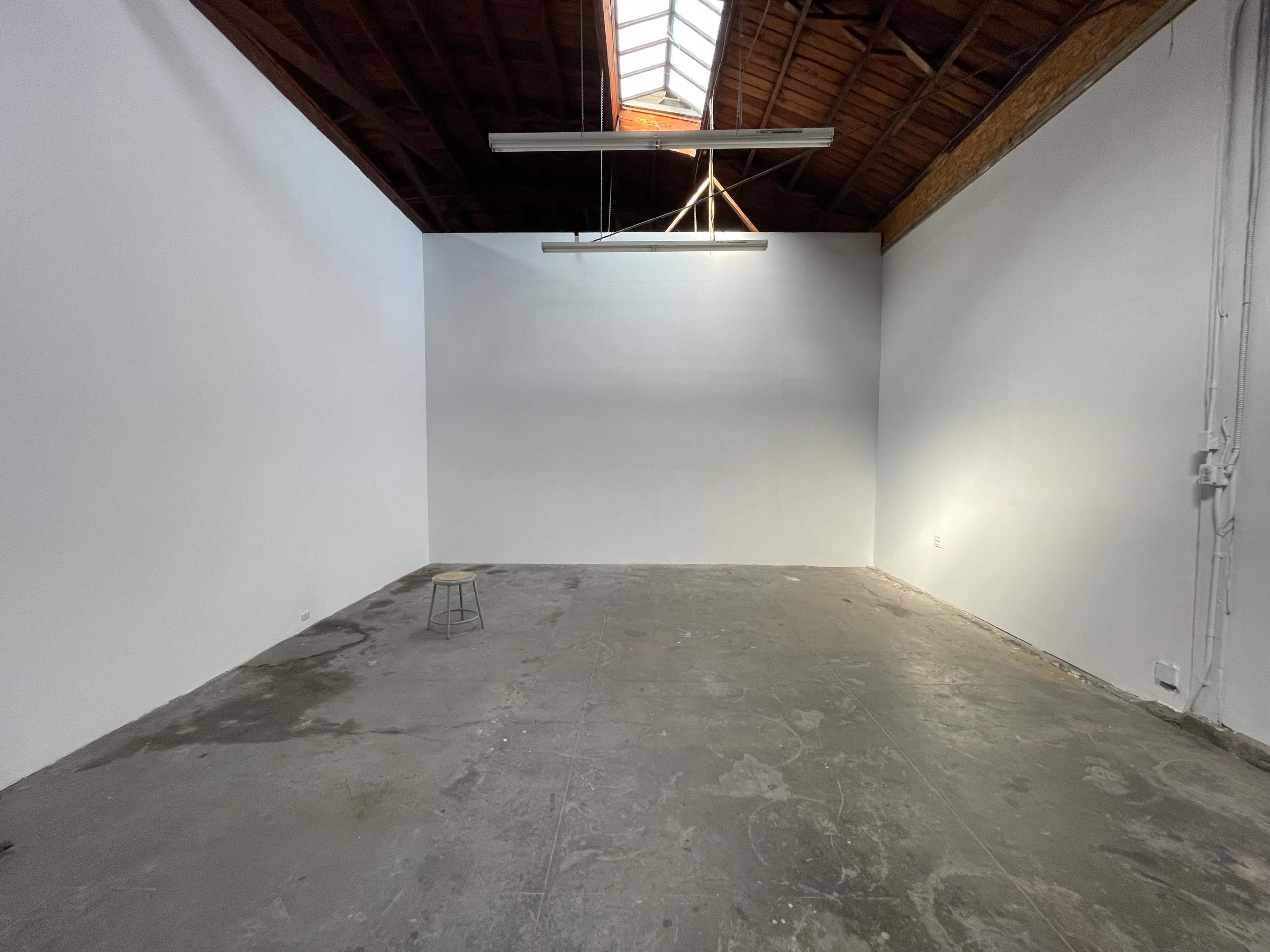 An empty gallery space features a concrete floor, white walls, and a light fixture hanging from the ceiling.