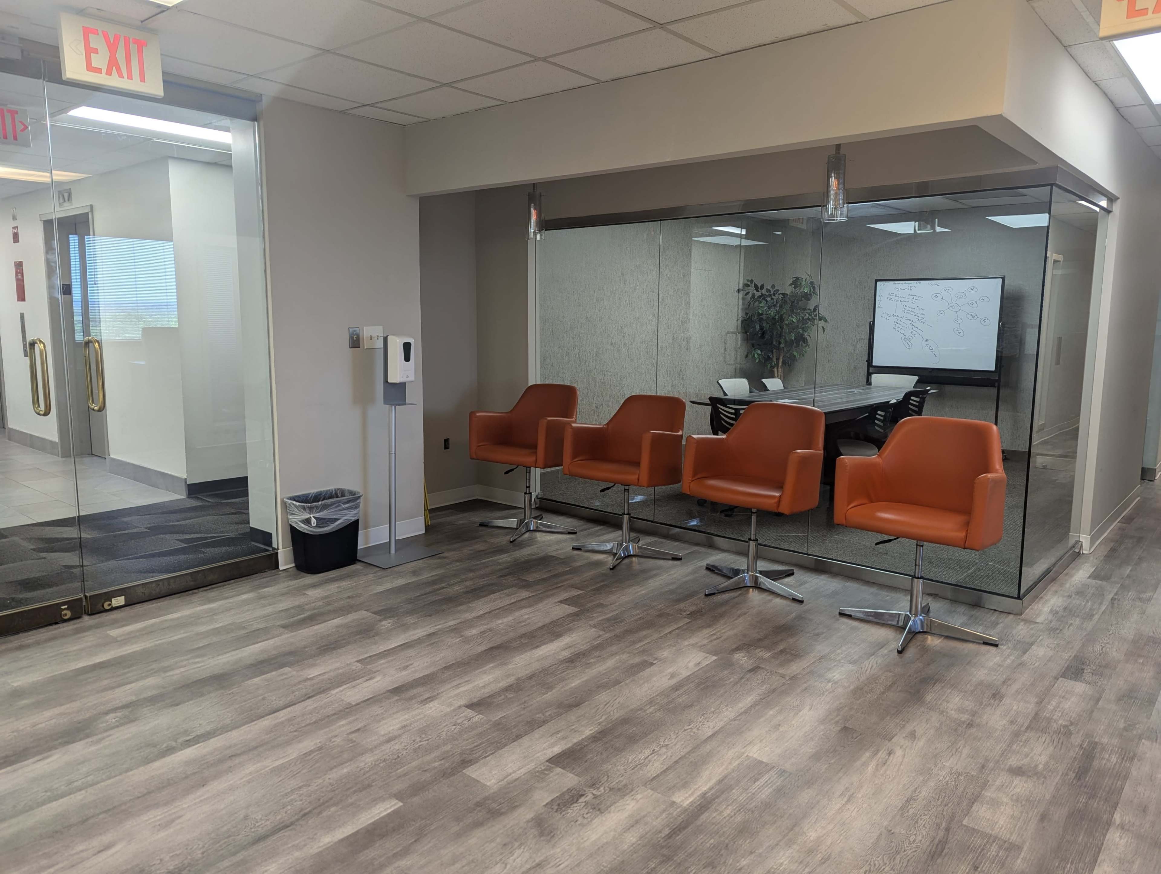 The image shows a modern office space with a glass-walled conference room and four orange chairs arranged in a waiting area.