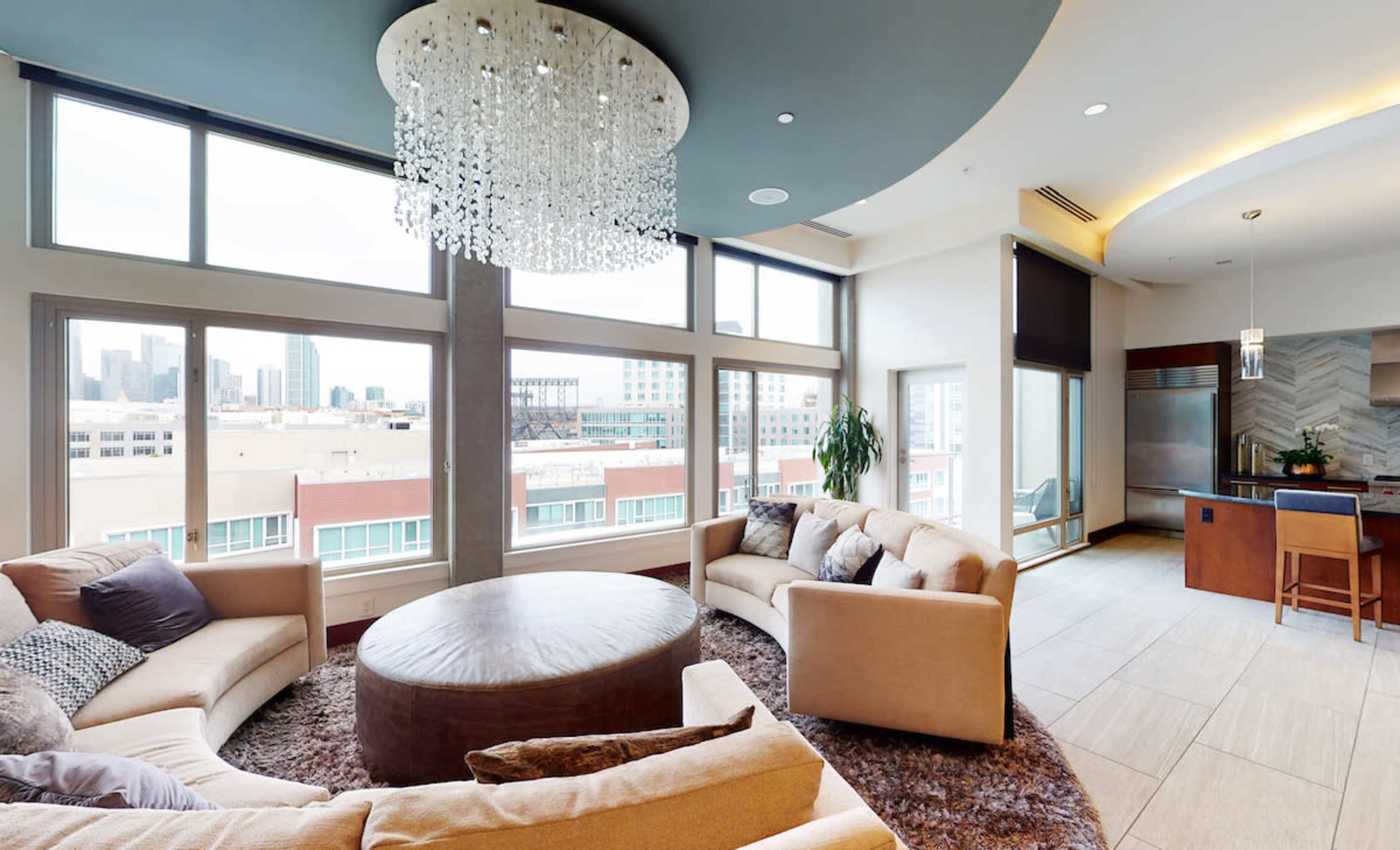 A modern living room with large windows, a round coffee table, and comfortable seating arranged around a plush area rug.