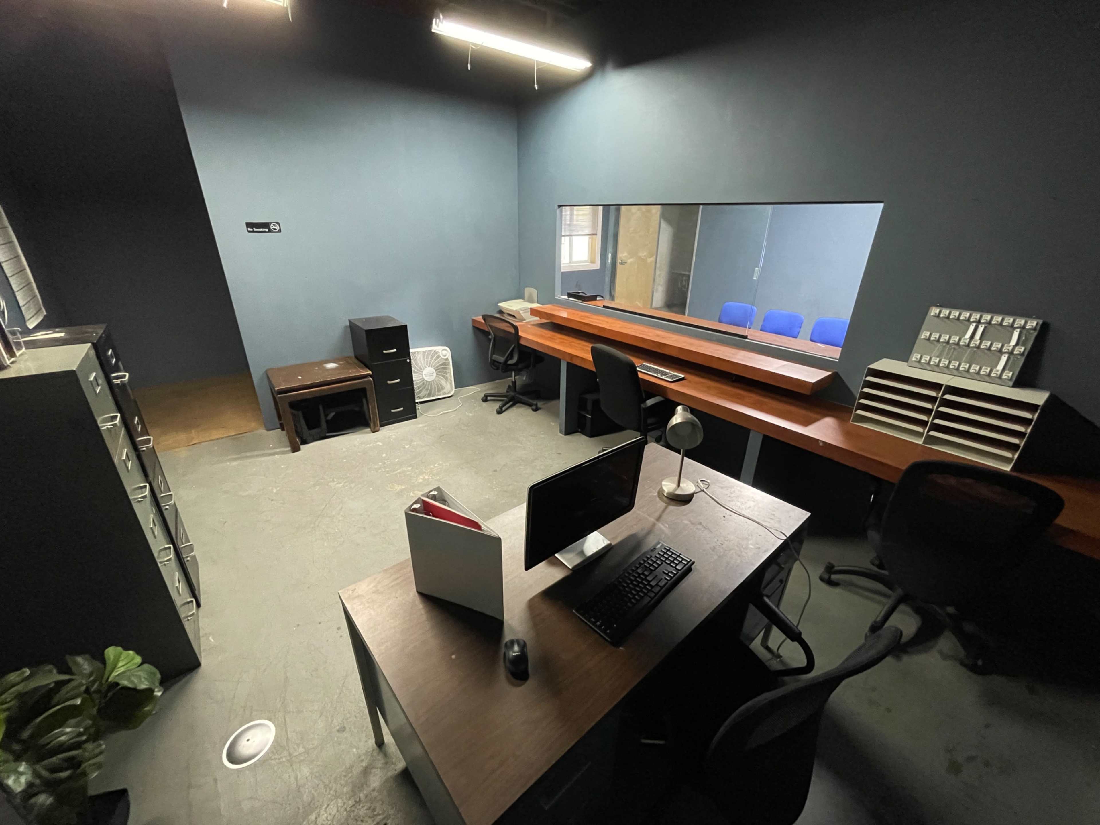 The image shows a sparsely furnished office with a desk, computer, filing cabinets, and a long table along the wall, all within a dimly lit space.