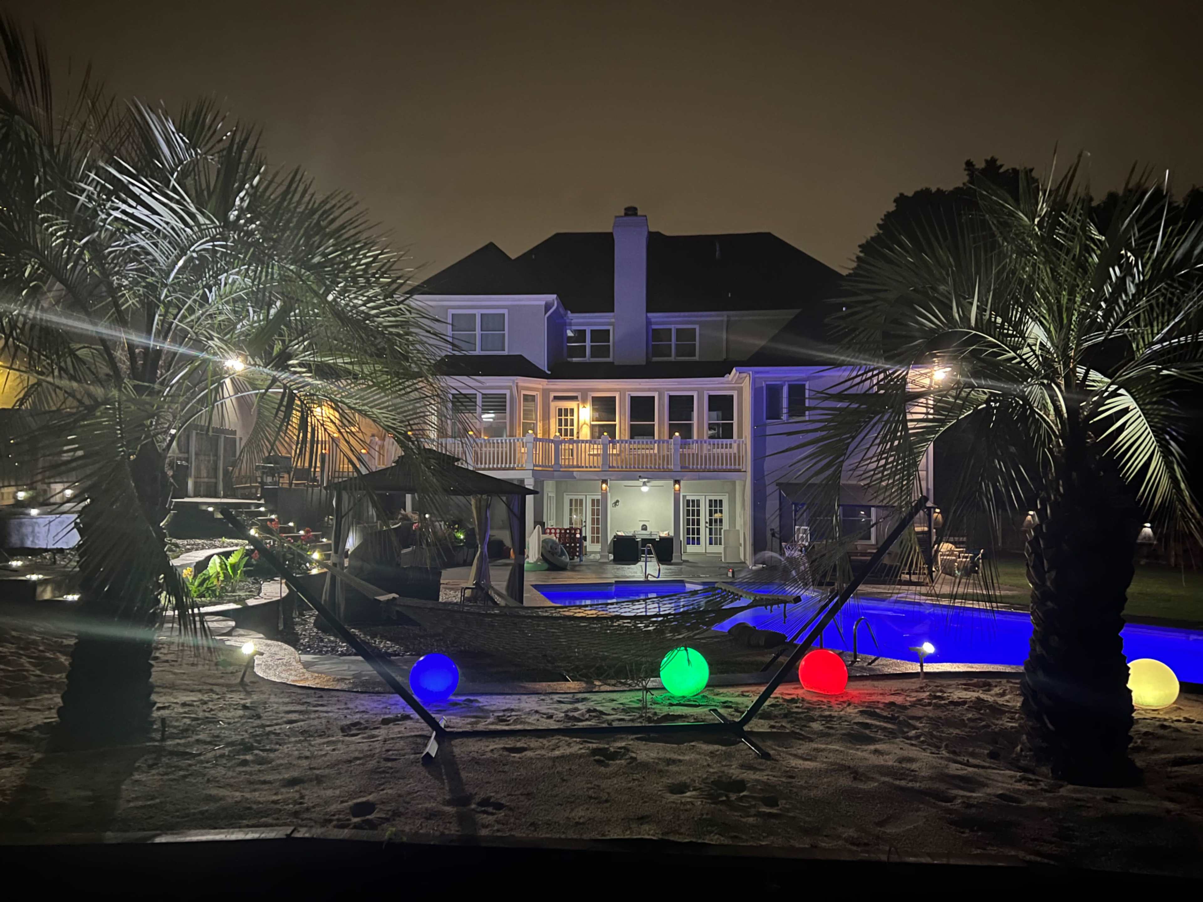 A modern house with palm trees and colorful lights surrounding a pool is illuminated at night.